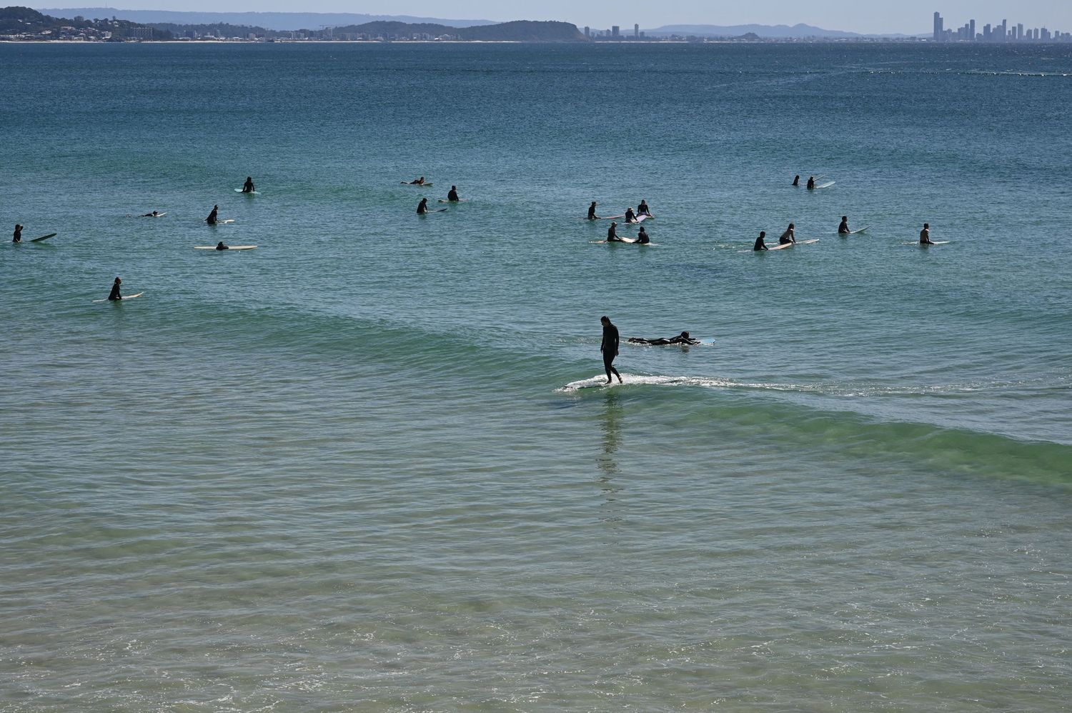 Surfers waiting for waves in turquoise ocean waters with a cityscape visible on the horizon on a sunny day.