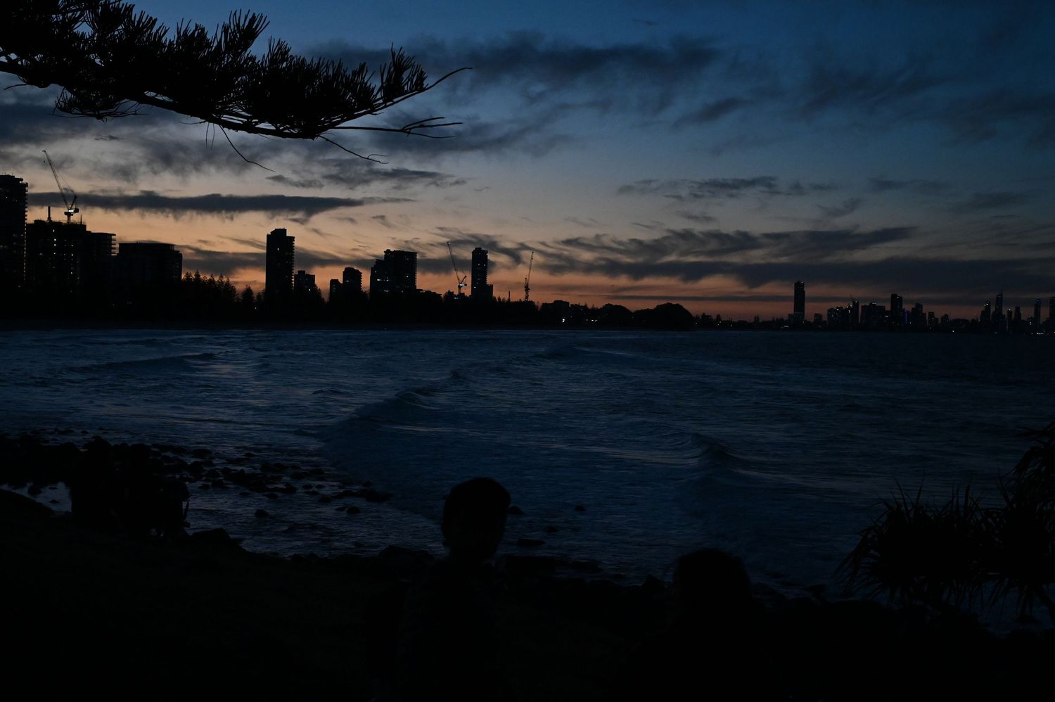 Dramatic dusk skyline silhouette over calm water with city buildings and colorful sunset sky reflected in twilight.