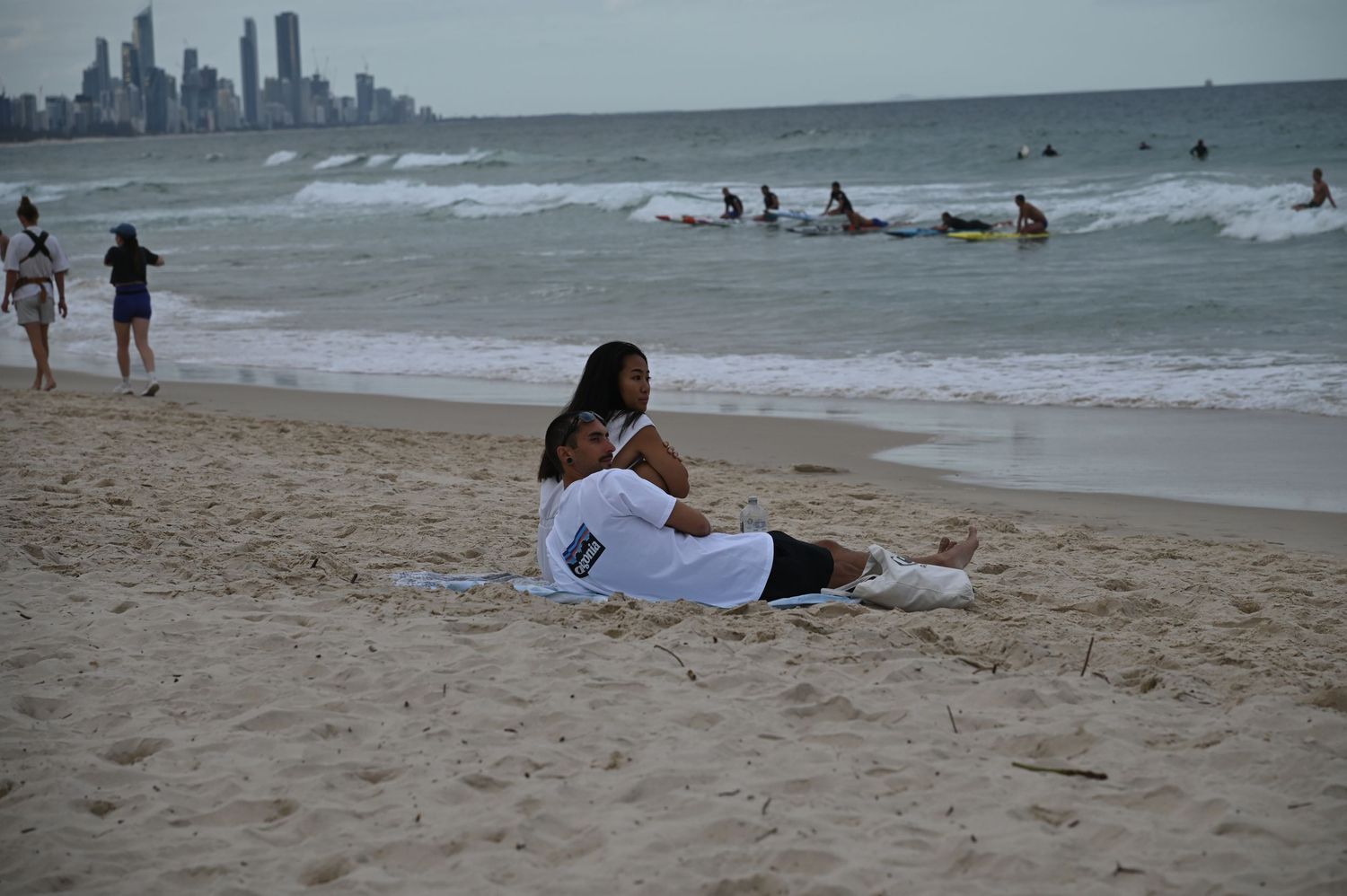 Couple relaxing on sandy beach with surfers in ocean waves and city skyline visible in background.