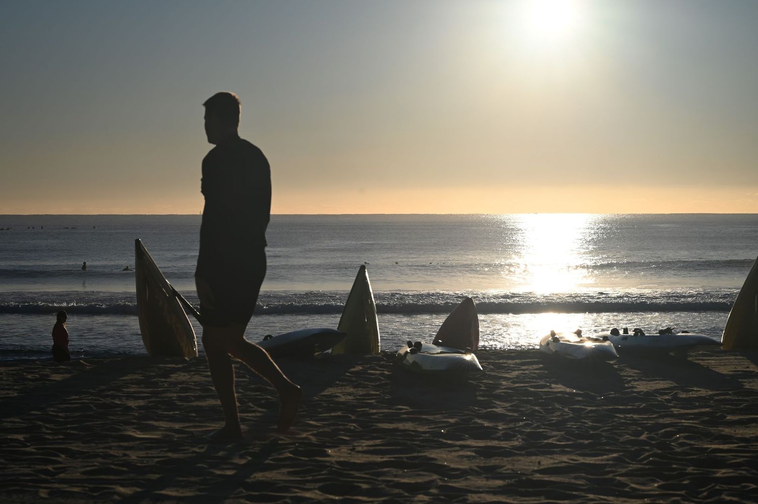 Silhouette of a man standing on a beach at sunset with surfboards in the sand and ocean waves in the background.
