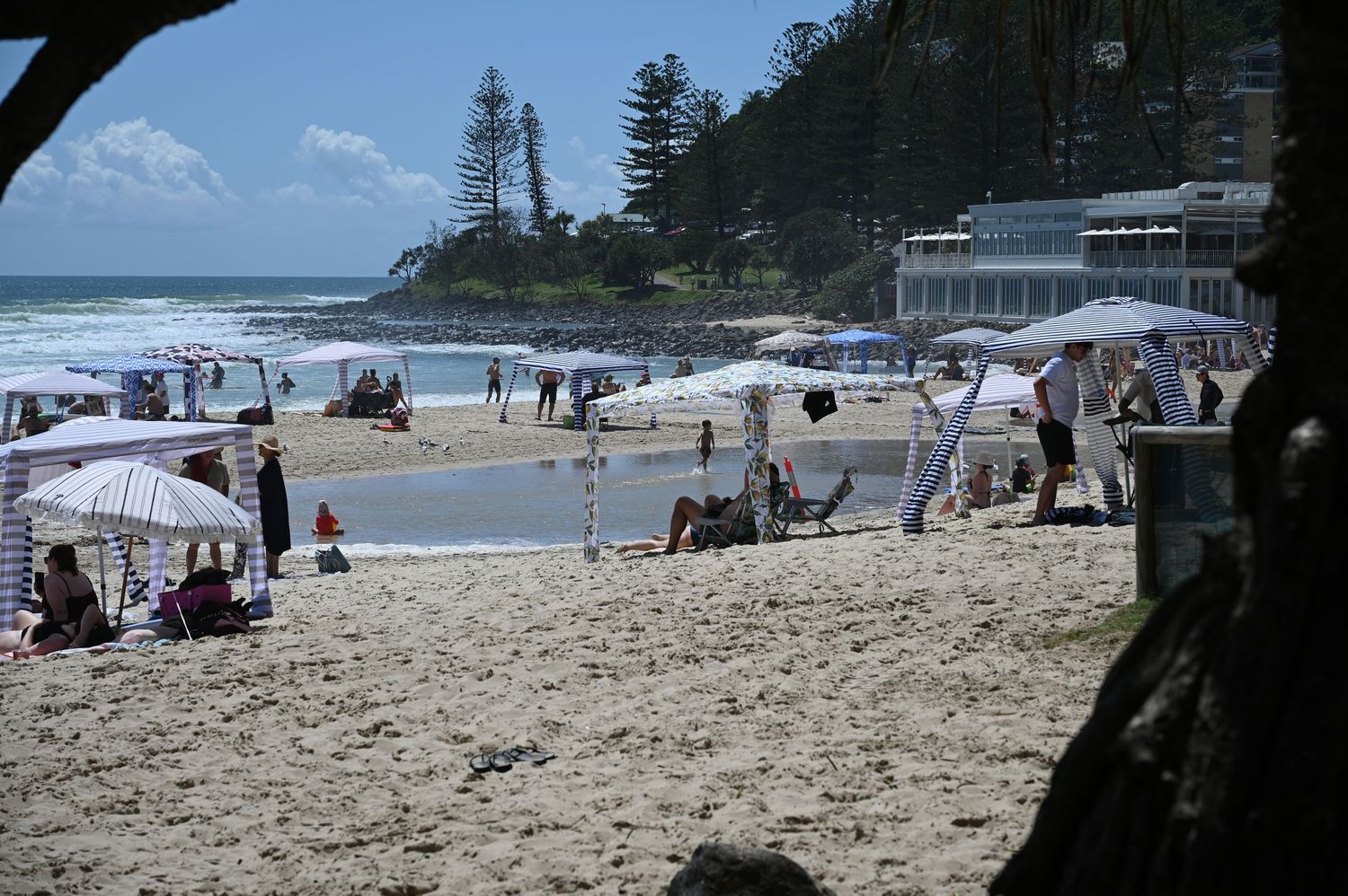 Busy Burleigh beach with white shade tents, beachgoers, and historic building near shoreline. Iconic Burleigh headland anchors the photo.
