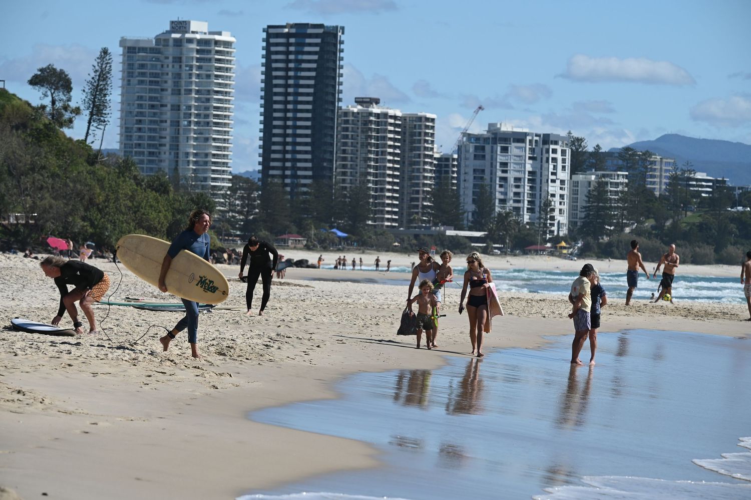 Beachgoers and surfers walk along a sunny Australian beach with high-rise buildings in the background. Golden sand. Blue seas. Relaxation.