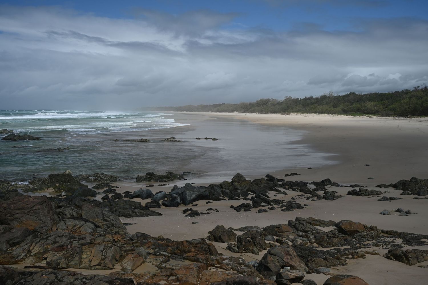 Rocky shoreline with sandy beach stretching into the distance under cloudy skies on an Australian coast.
