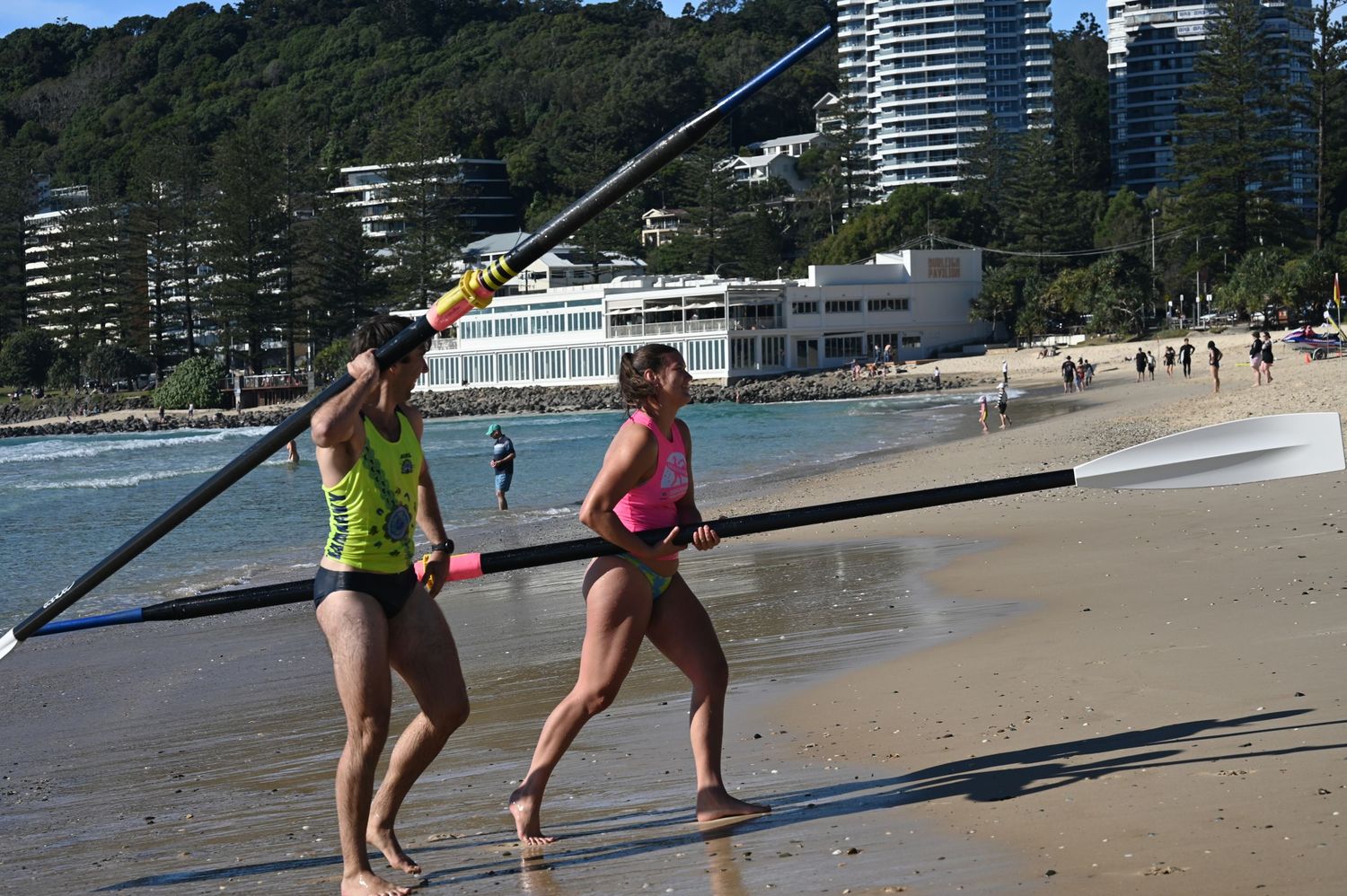 Two women in swimsuits carry a long oar on a sandy beach with ocean and buildings in the background.