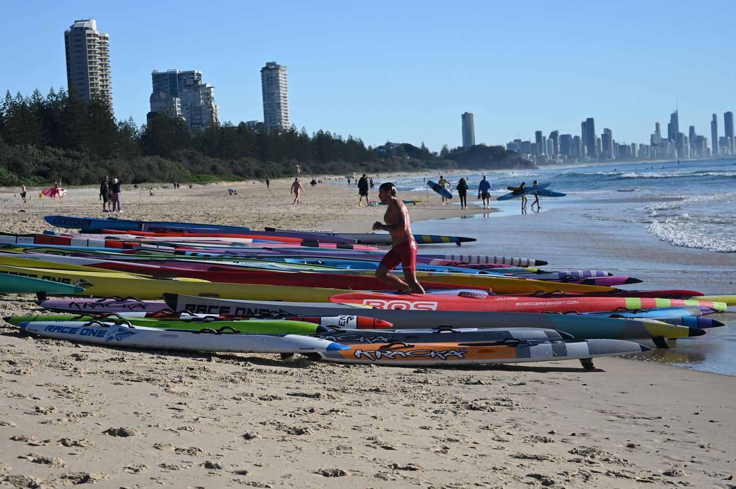 Colorful surf ski kayaks lined up on sandy beach with surfers and Gold Coast skyscraper skyline in background.