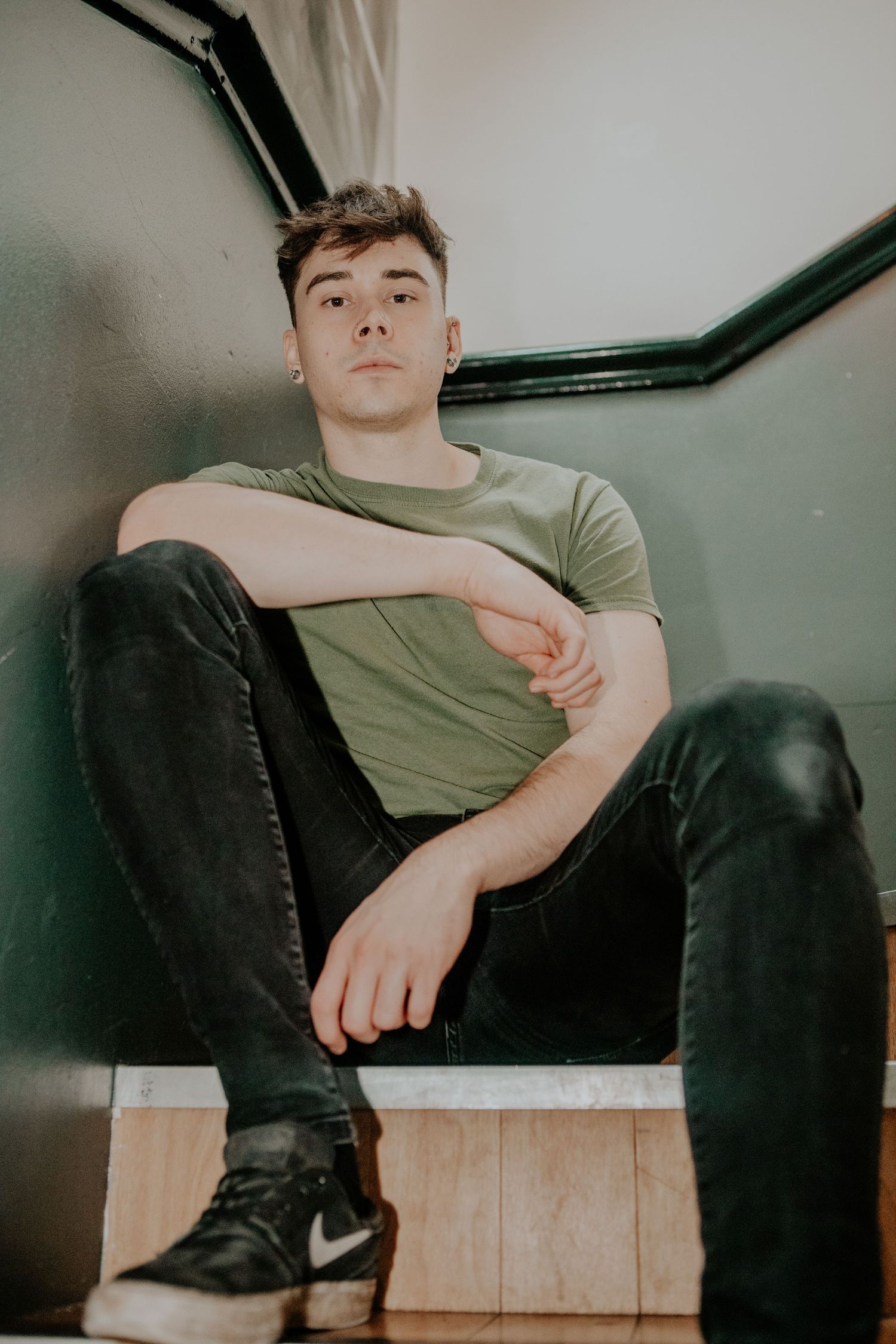 Dissonants drummer in olive t-shirt and black jeans sitting casually against grey wall.