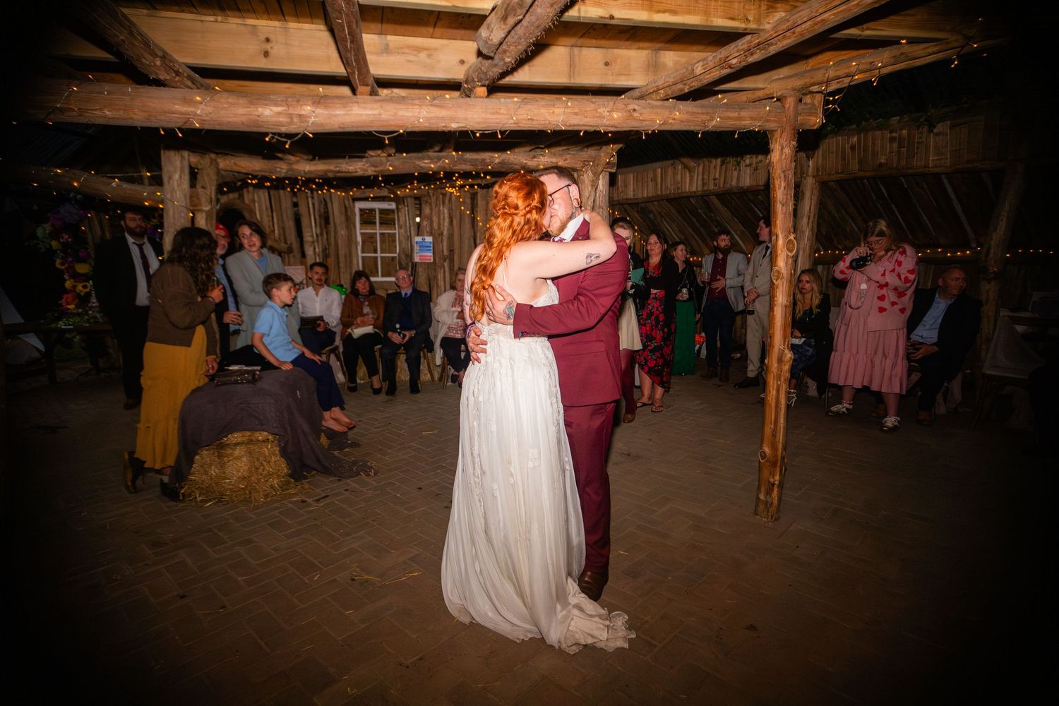 A couple in burgundy suit and white wedding dress share their first dance in a rustic wooden barn venue.