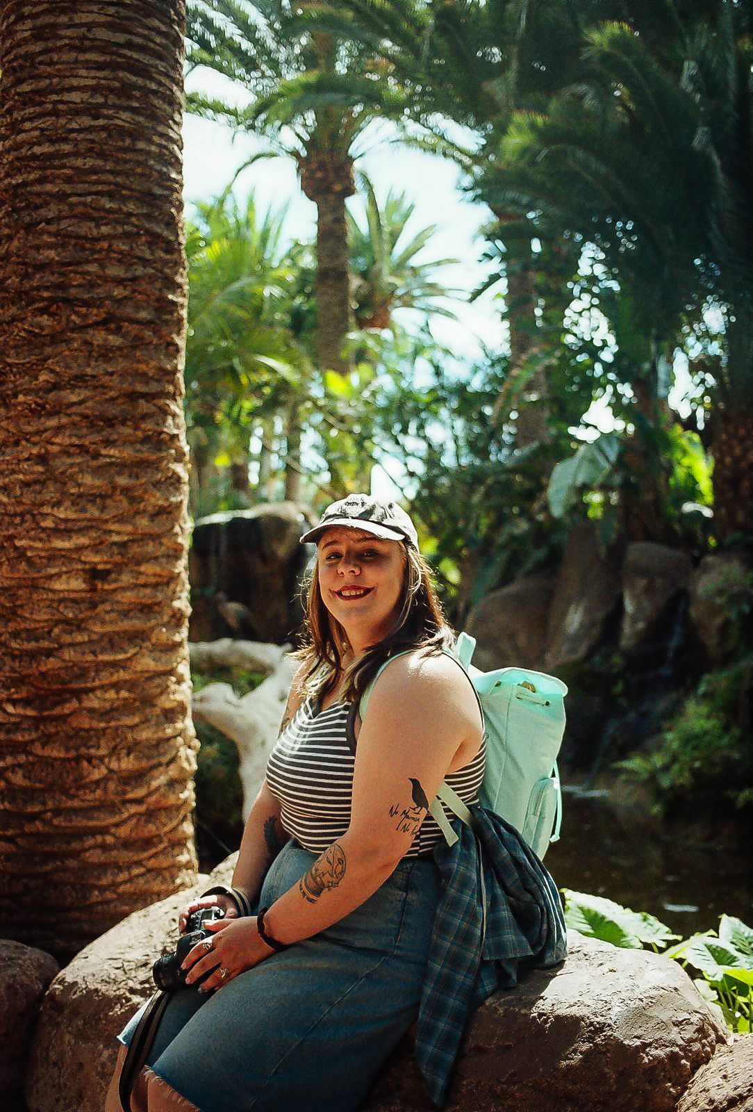 Emily wearing a striped top and backpack sits on a rock among palm trees in a tropical setting.