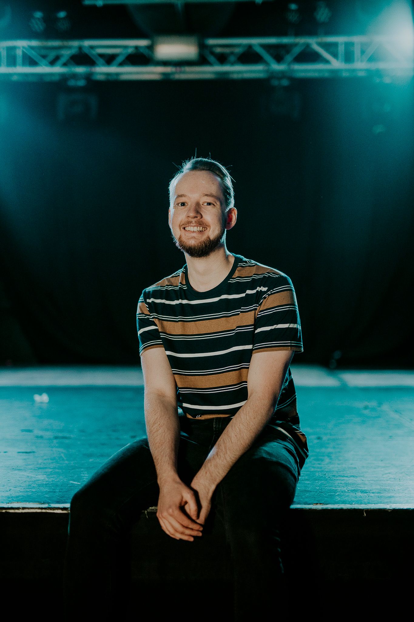 Dissonants bassist in striped shirt sitting on stage under blue theatrical lighting.