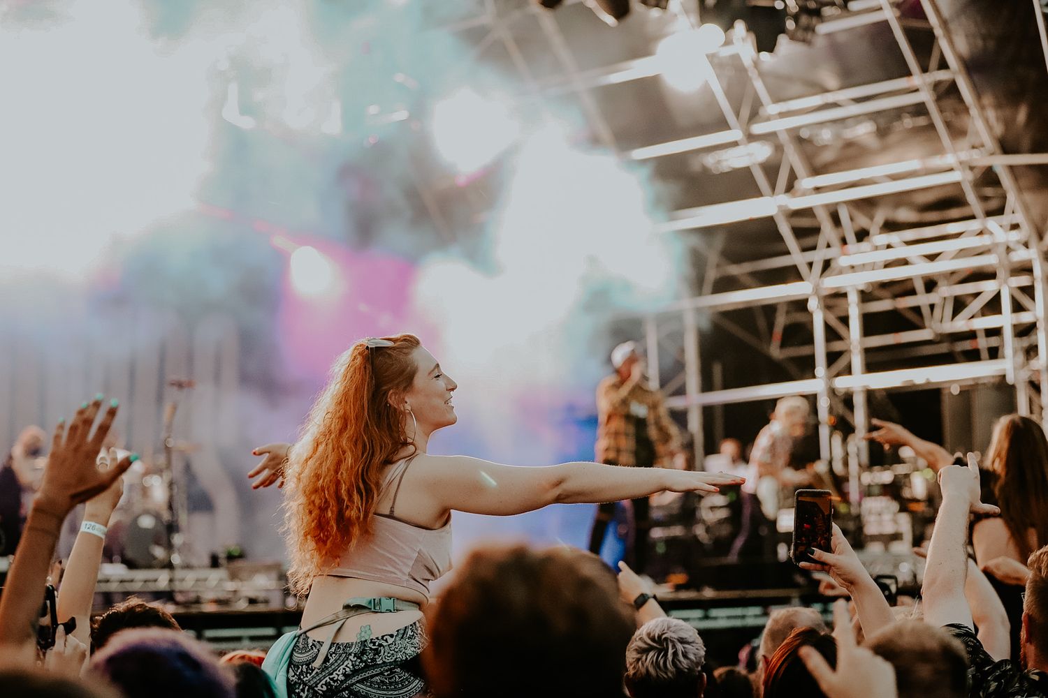 Concert crowd enjoying music festival with smoke effects and colorful lights while dancing at outdoor venue.