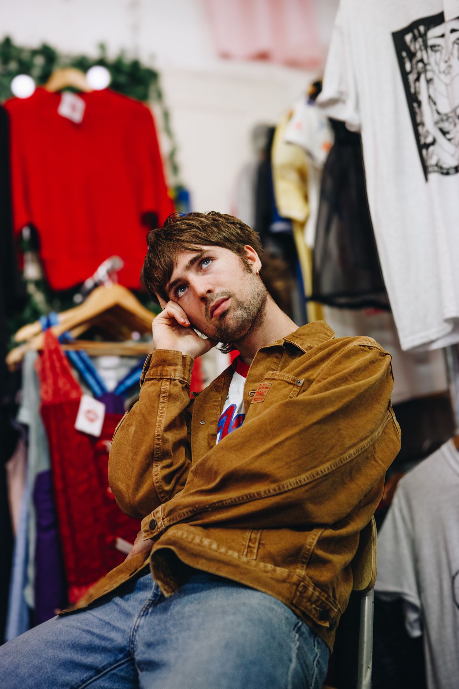 Will Joseph Cook is in a brown jacket, leaning on their hand while sitting among clothing racks.