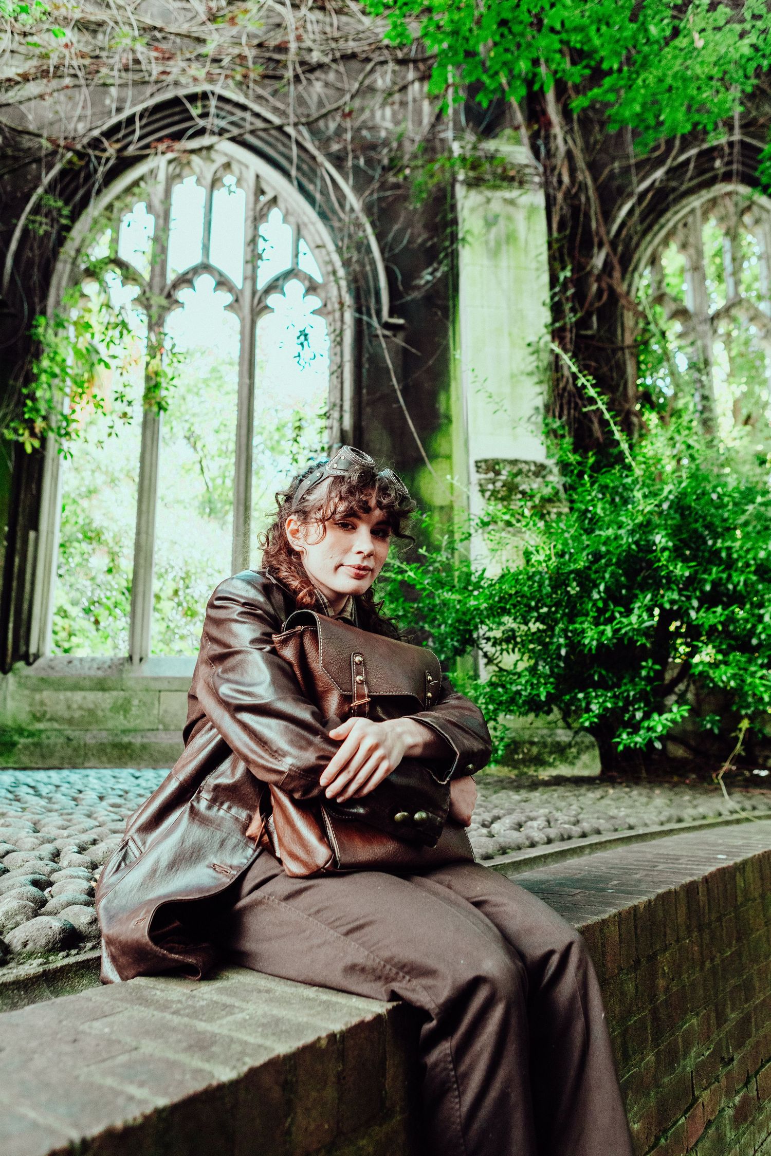 A person in brown leather attire sits on stone architecture surrounded by gothic windows and greenery.