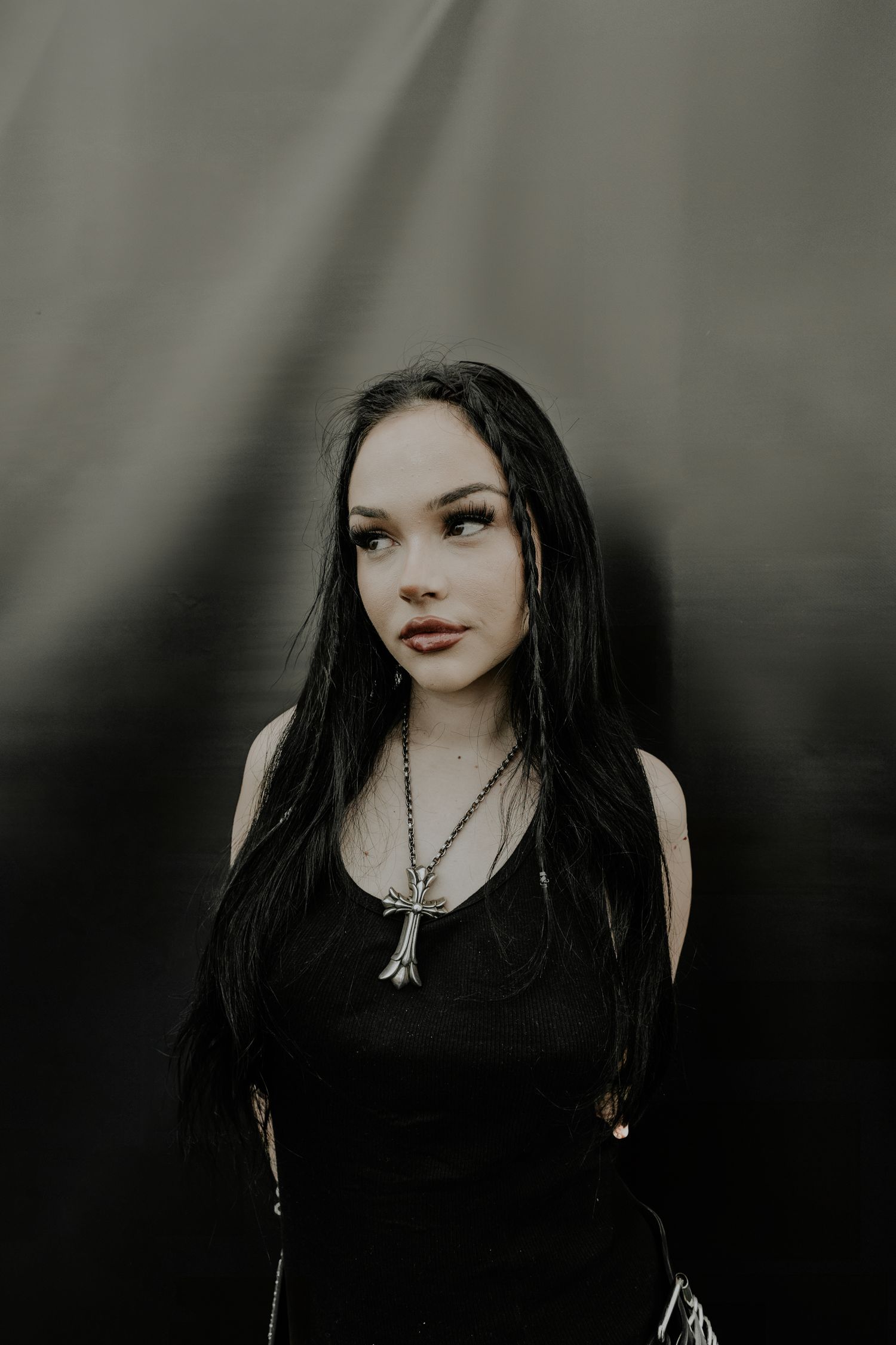 Maggie Lindemann in black attire with long dark hair and a pendant necklace against a grey background at Slam Dunk Festival.