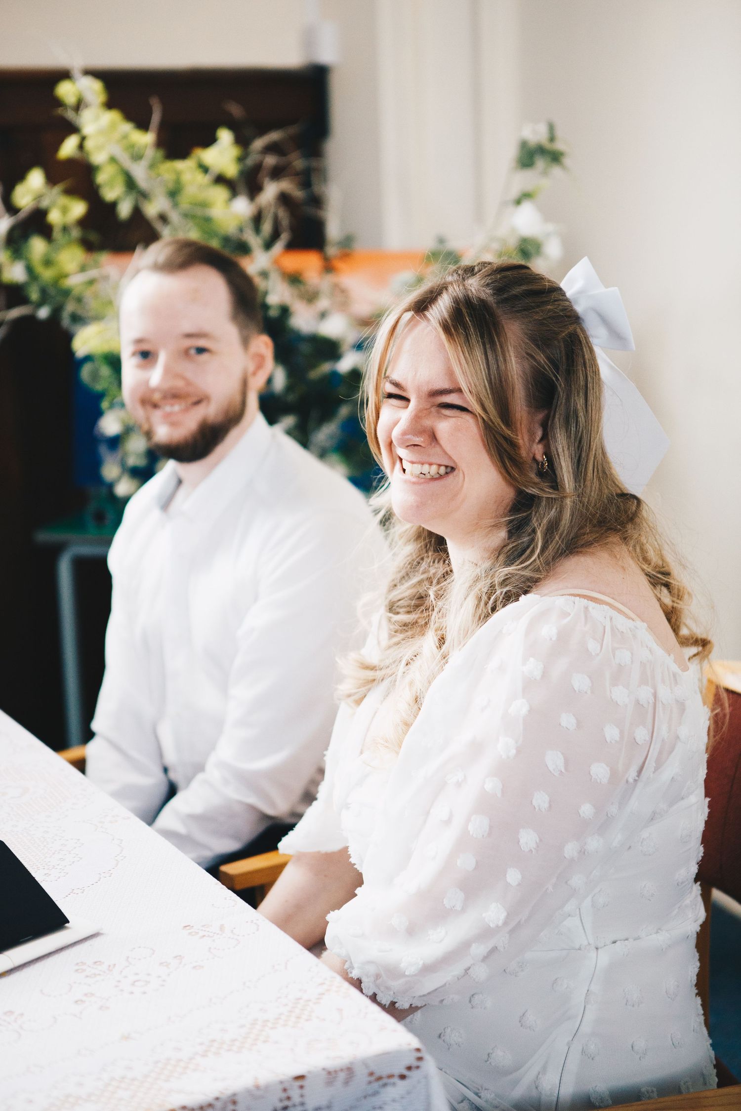 Couple sits together at a piano in white wedding attire while laughing joyfully during a candid moment.