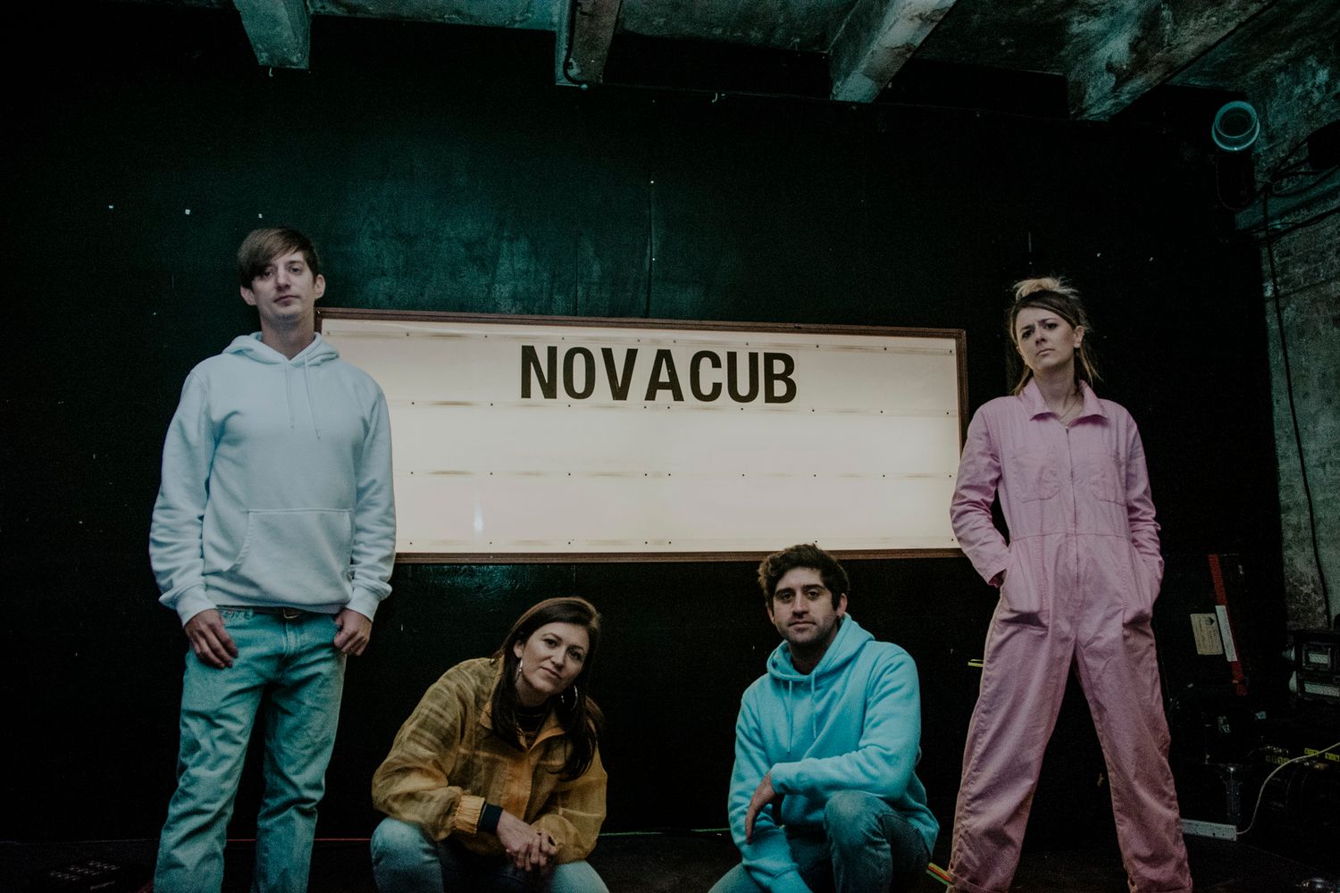Band members posing casually in front of NOVACUB sign in dark indoor setting.
