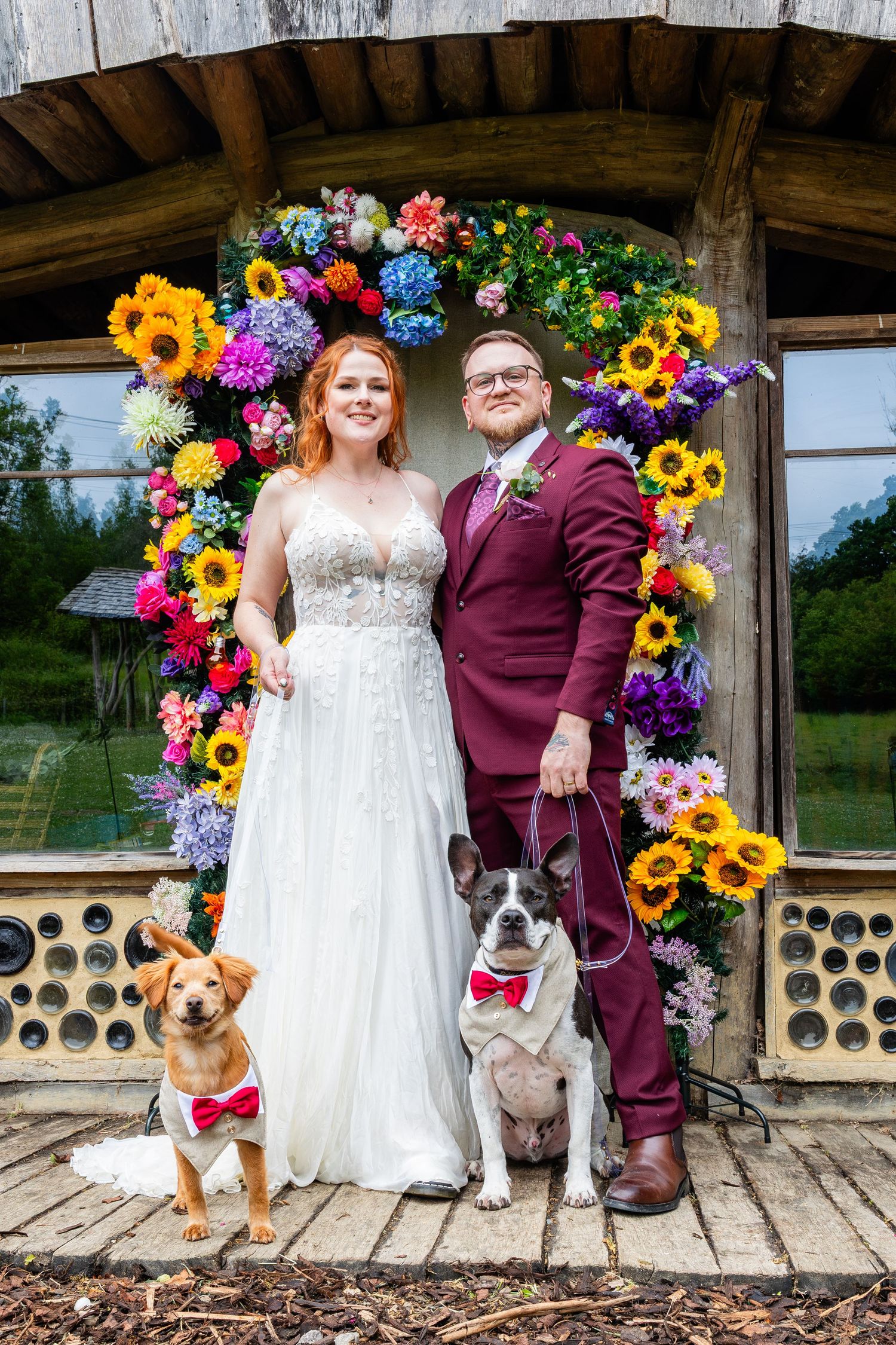 A couple in wedding attire poses under a vibrant floral arch with their two dogs wearing red bowties.