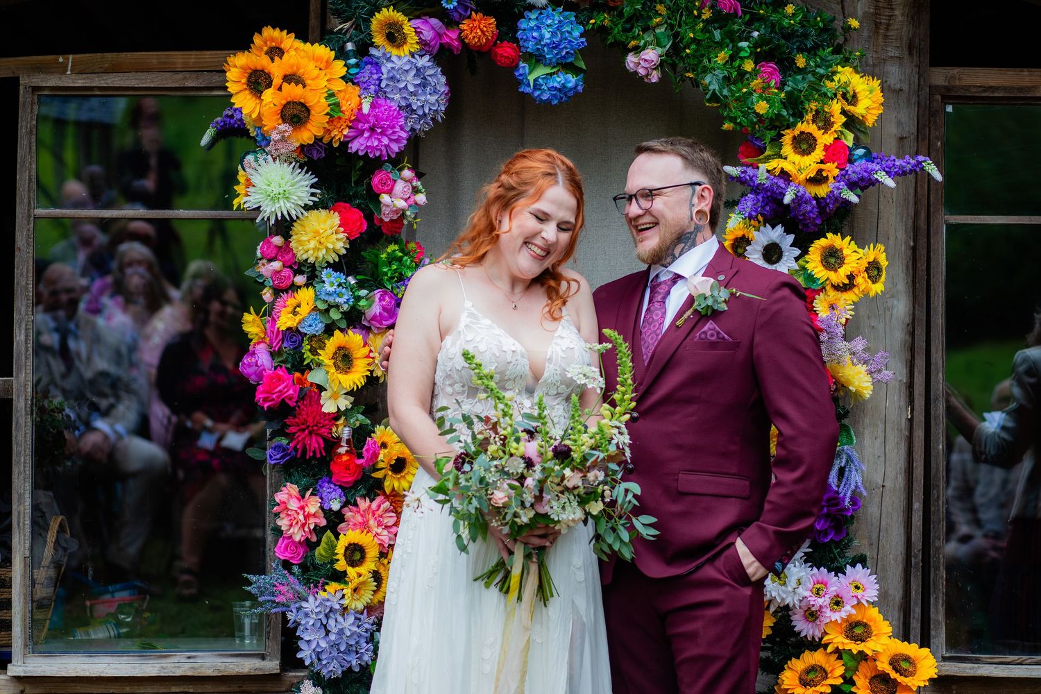 A wedding couple poses under a vibrant floral arch with sunflowers, purple and blue blooms against a glass wall.