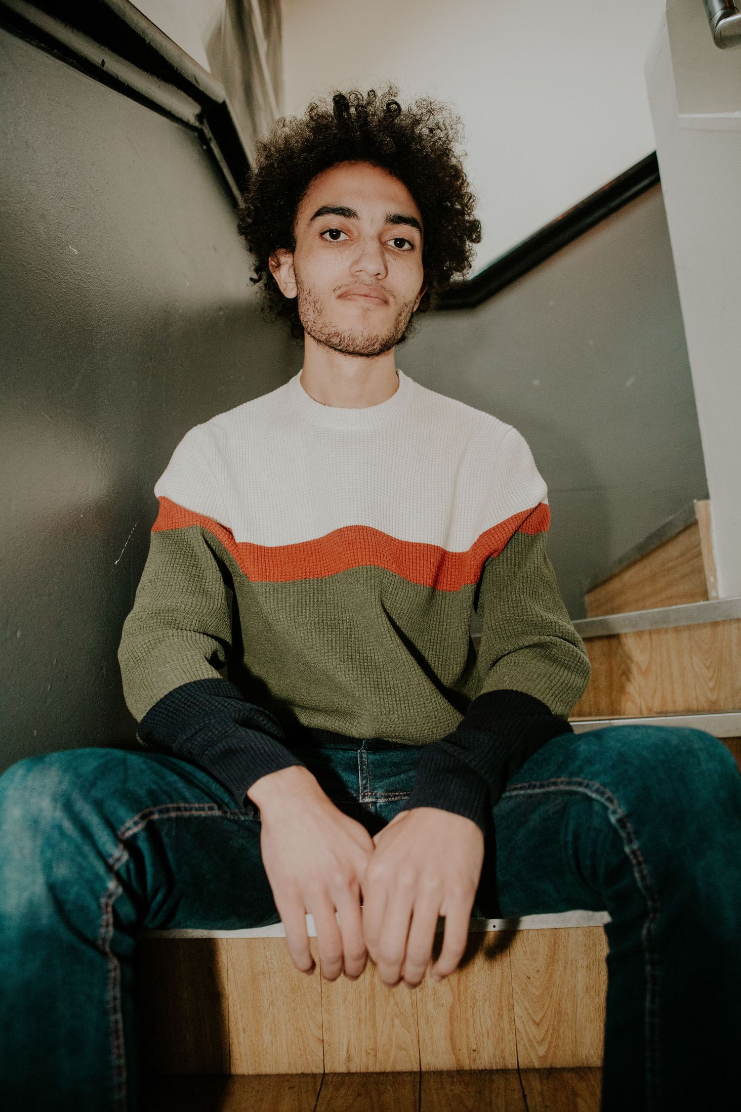 Dissonants ex-drummer sitting on wooden steps wearing colorblock sweater.