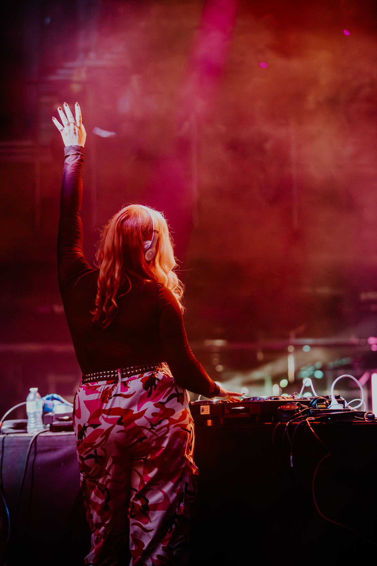 DJ performs at a concert with raised hand against red smoky atmosphere in colorful patterned pants.