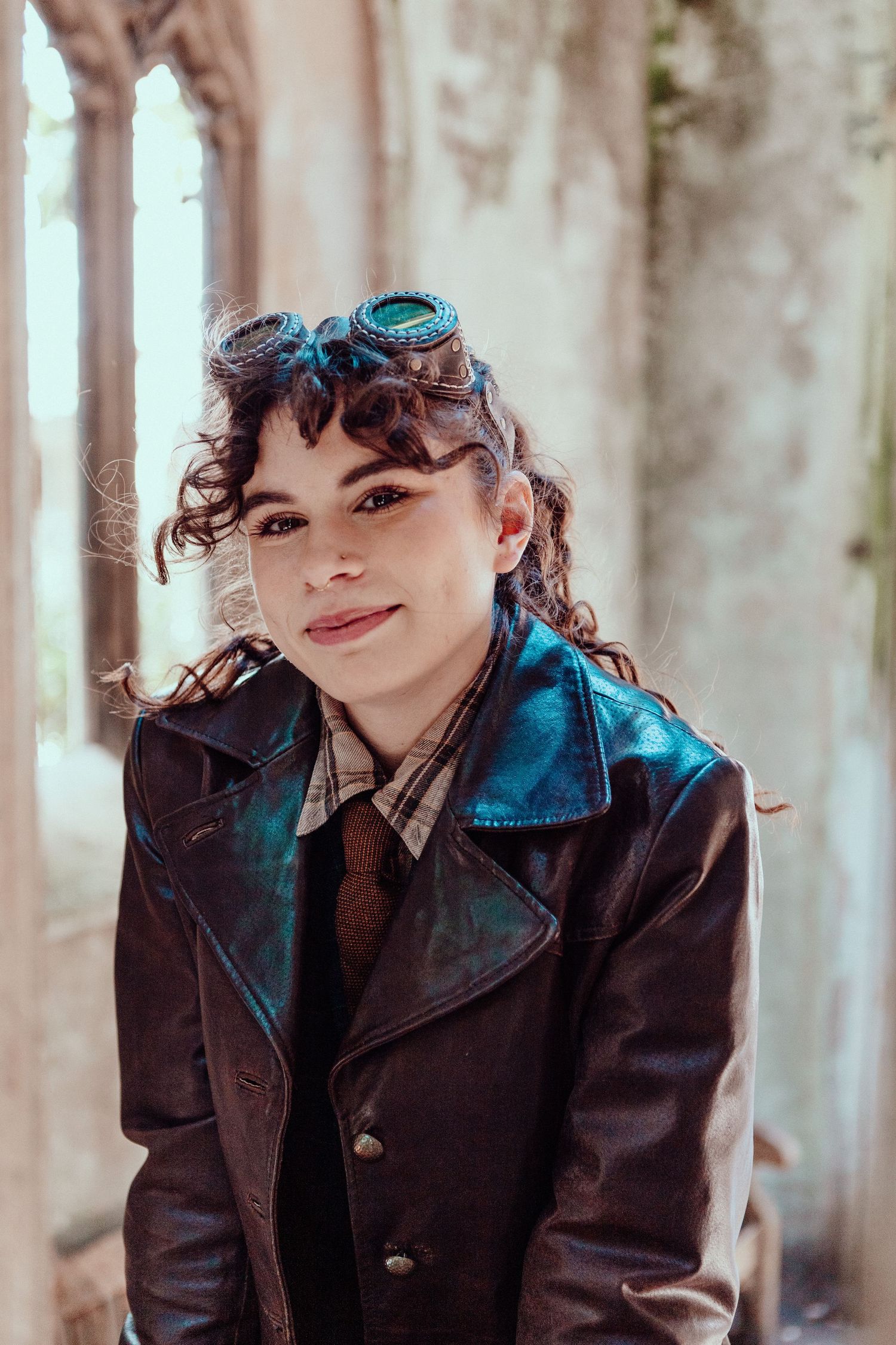 A person in a leather jacket and tie poses near gothic architecture with sunglasses on their head.
