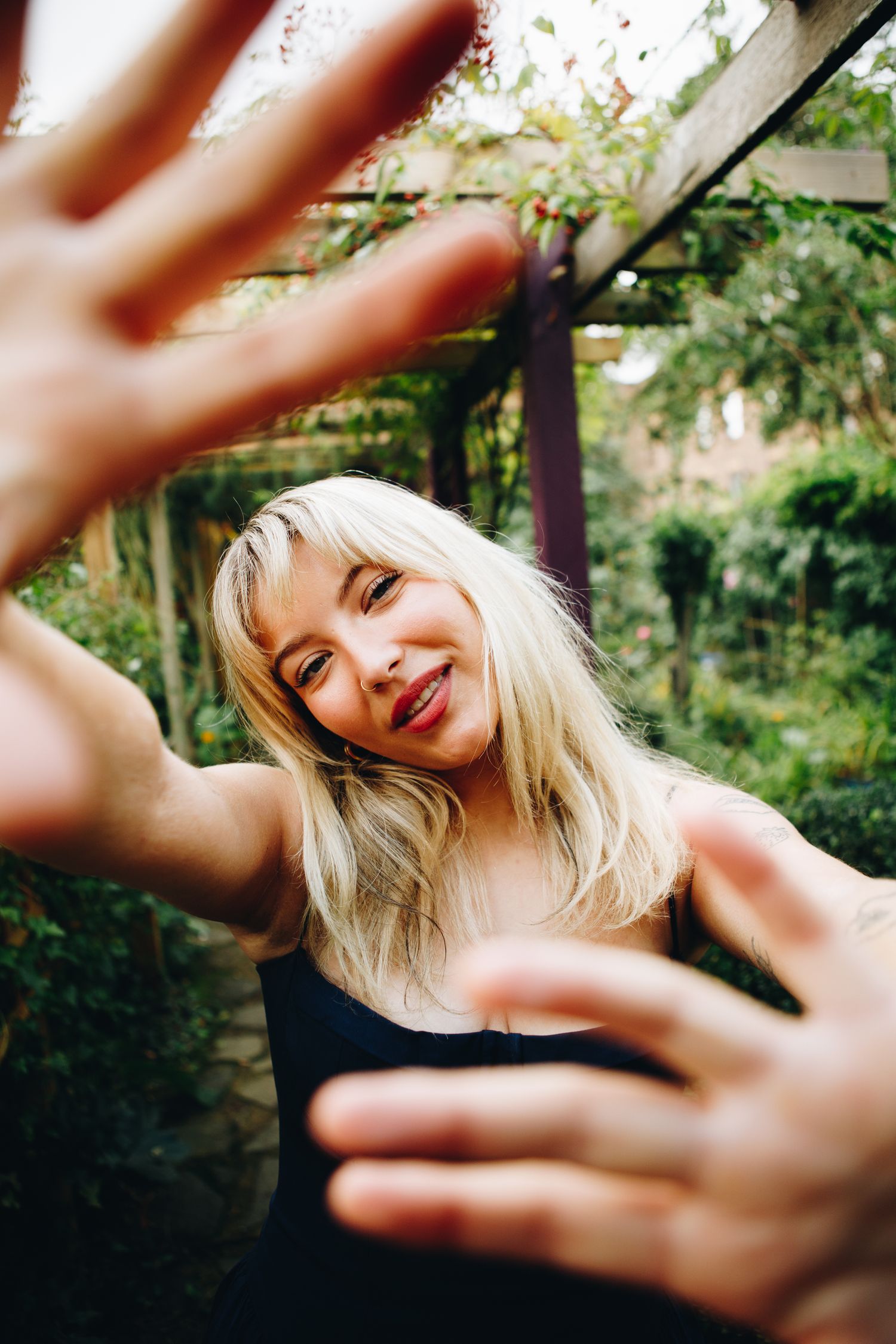 A playful outdoor portrait shot of Cate Canning through reaching hands in the foreground with natural garden scenery.