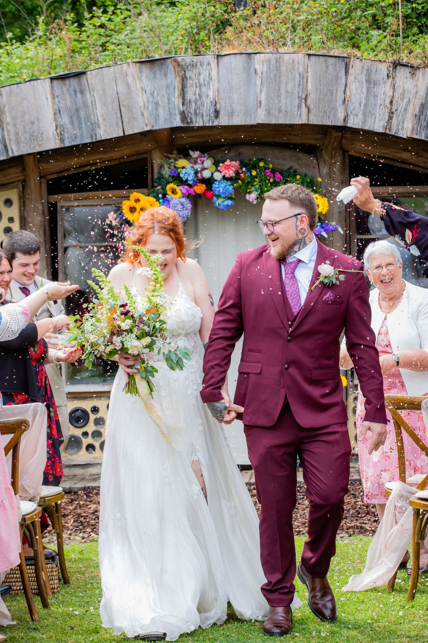 A joyful wedding scene in a garden with a burgundy-suited couple walking together beneath a rustic wooden arch.