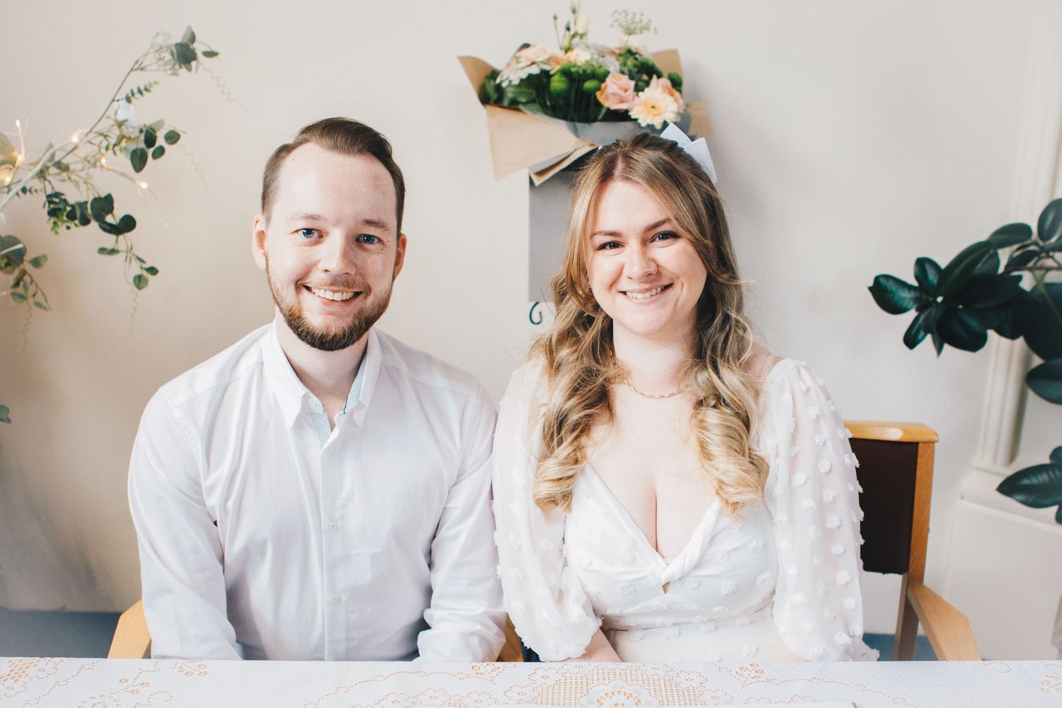 Two people sitting together at a table, wearing white, with greenery decorations on the wall behind them.