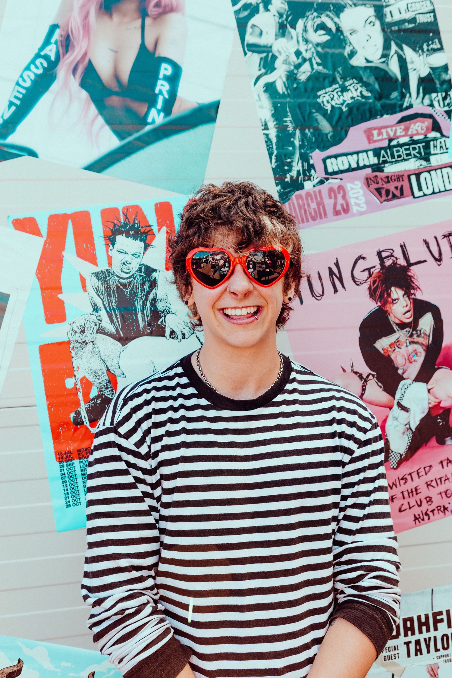 Noahfinnce in a striped shirt and sunglasses poses in front of a wall covered in music posters.