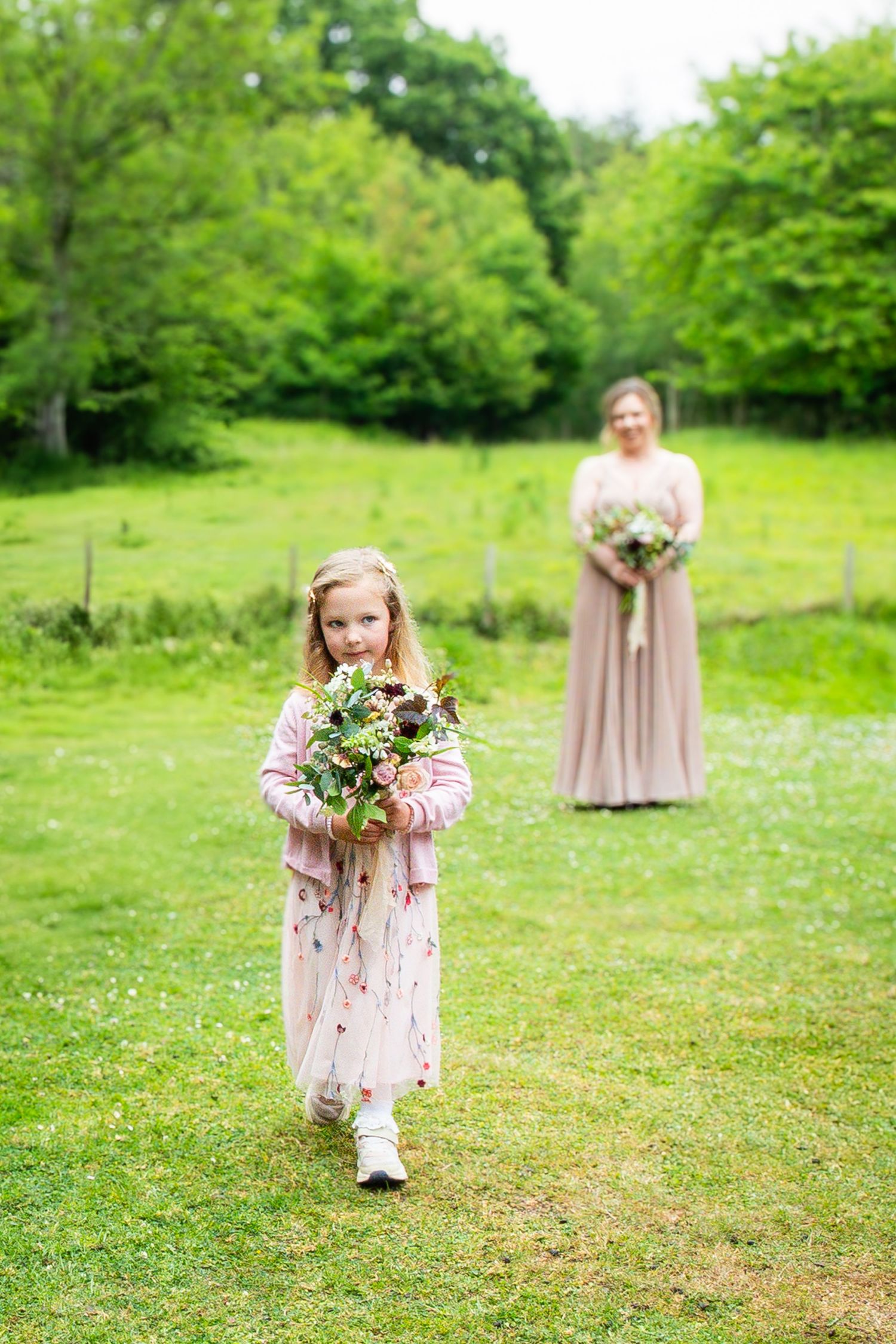 Young flower girl in a floral dress walks through a green garden meadow holding a spring bouquet.