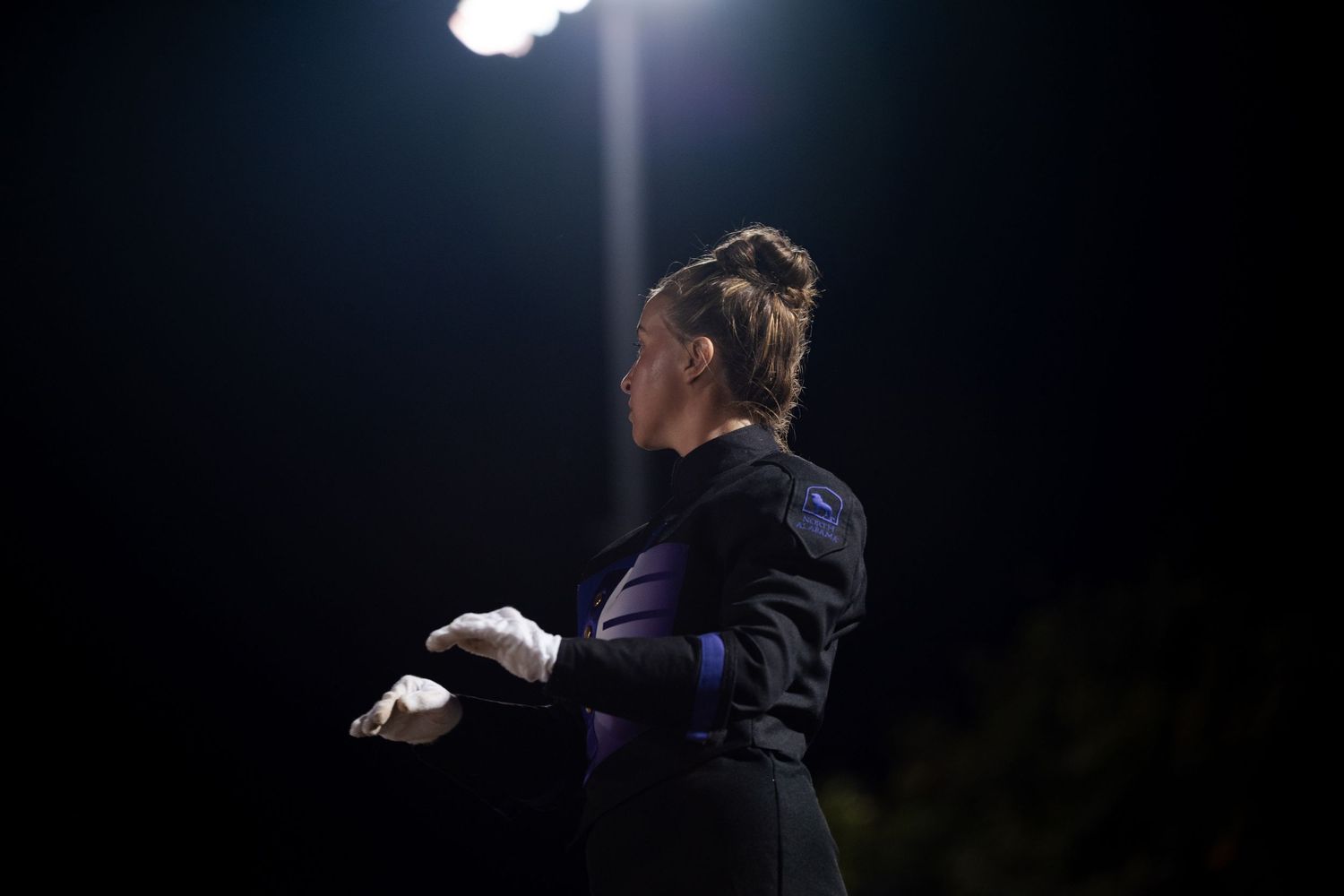 Female marching band member in black uniform holding equipment under a spotlight at night.