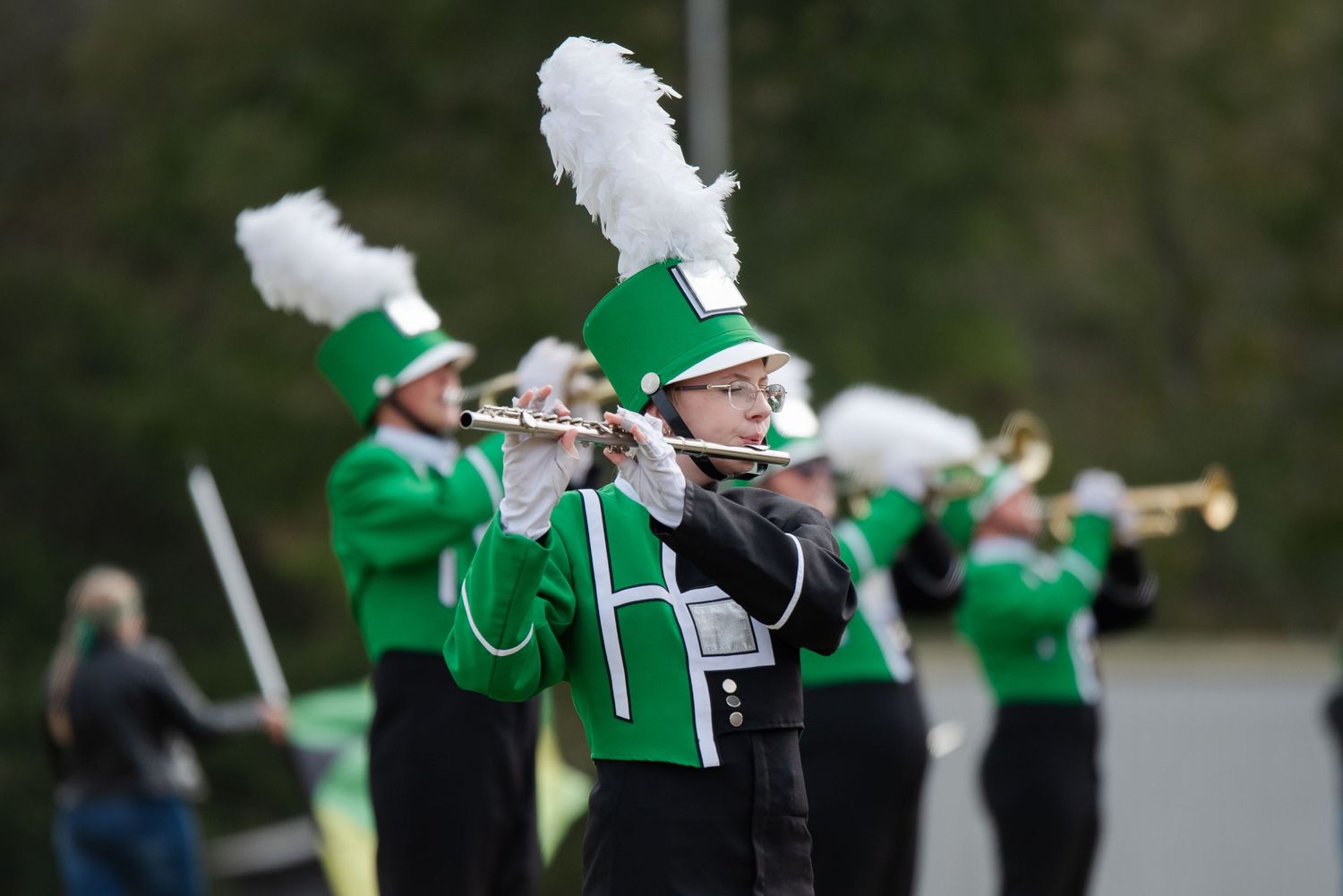 Green-uniformed marching band members play brass instruments during an outdoor performance.