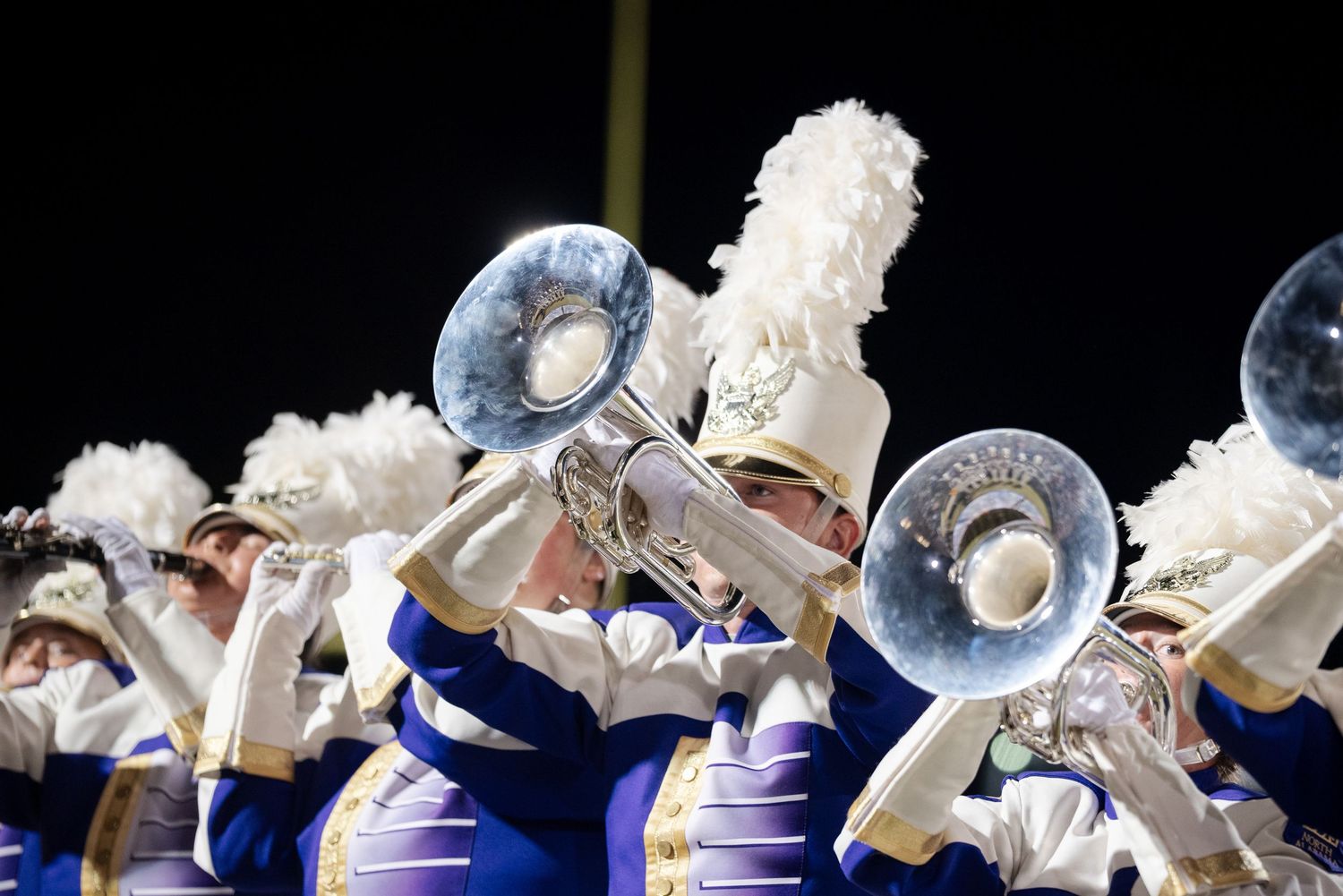 Marching band members in purple and gold uniforms play brass instruments at night.