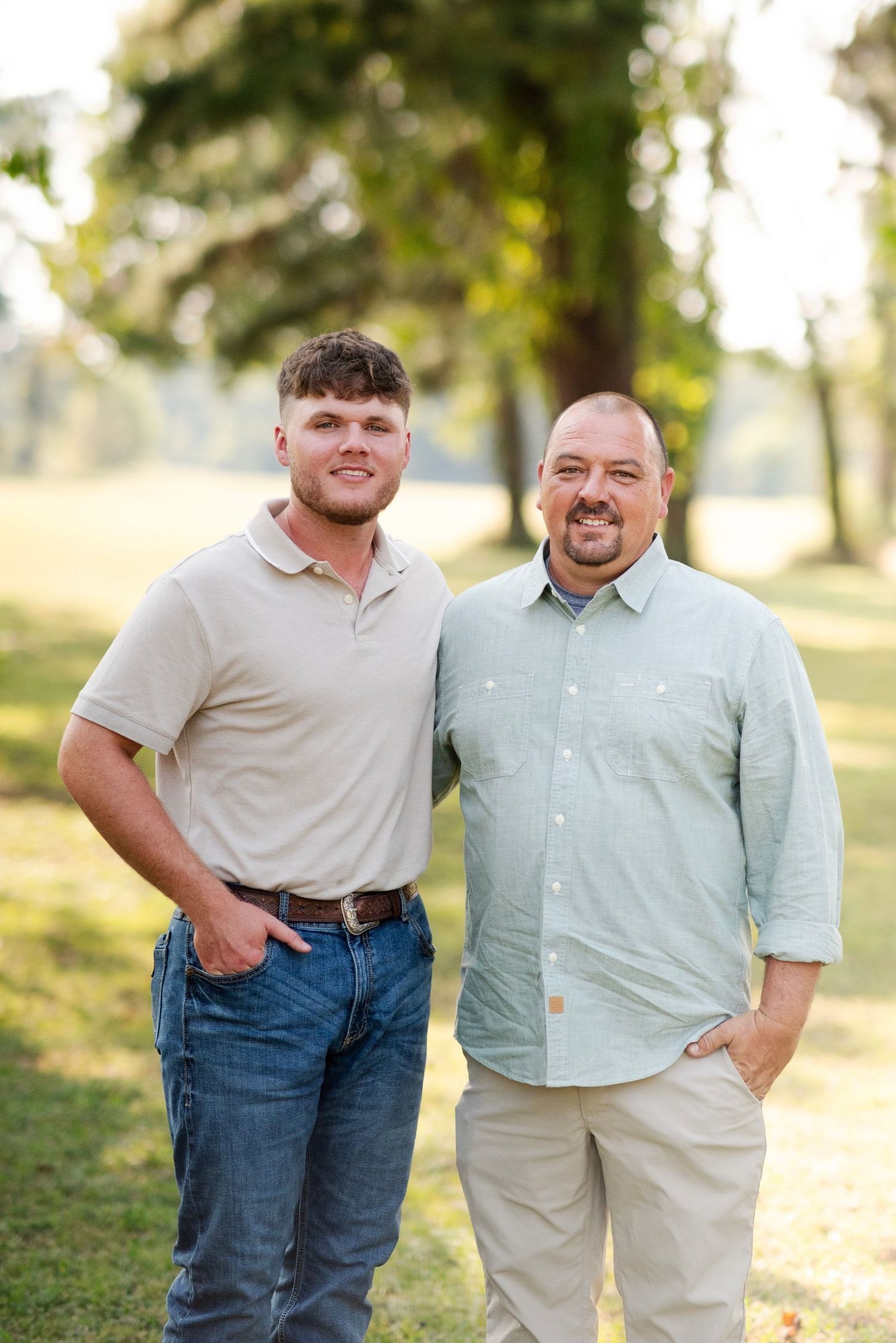 Two men, one young and one middle-aged, smile together in a sunlit green park setting.