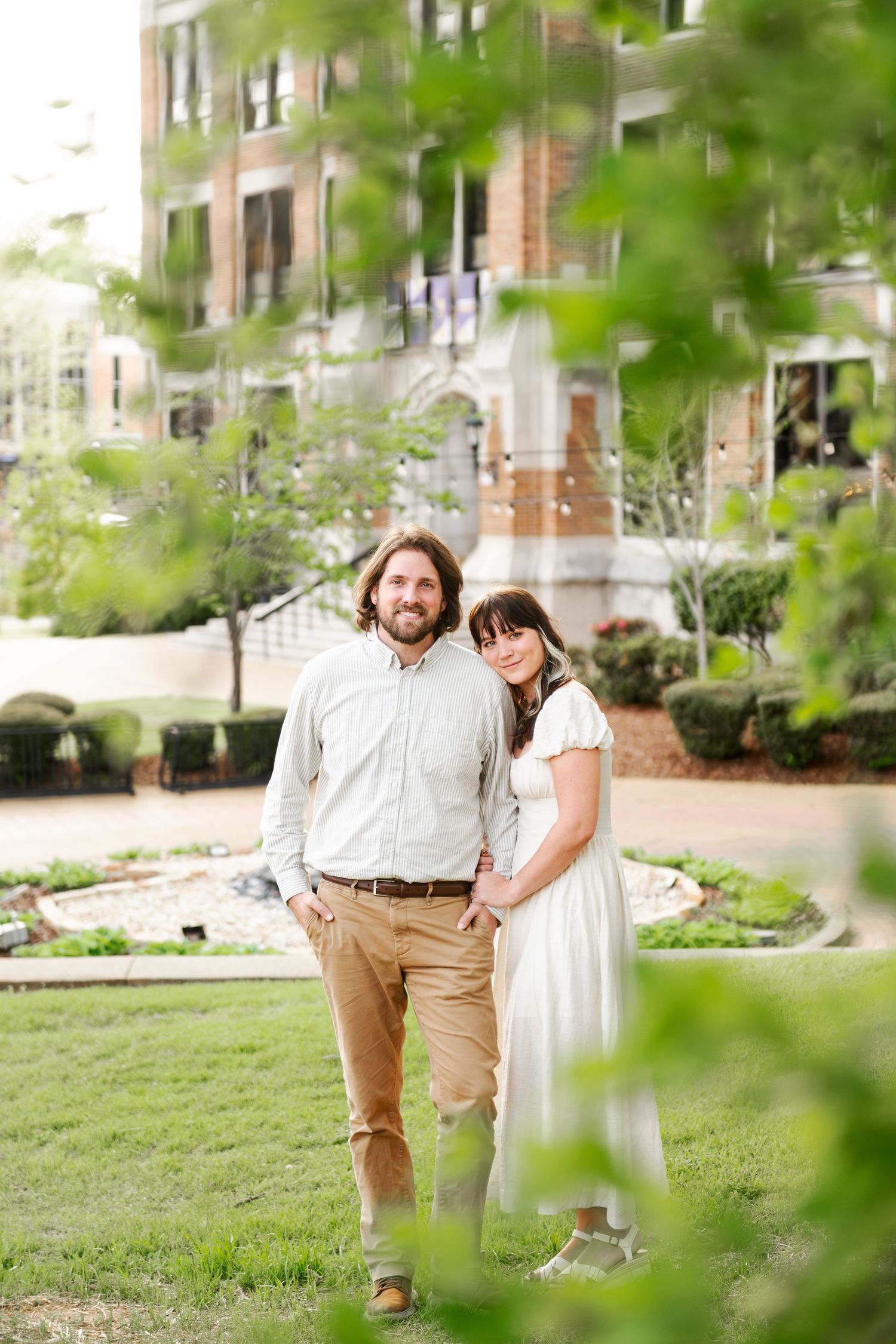 A couple stands together smiling in a lush green courtyard outside a brick university building.