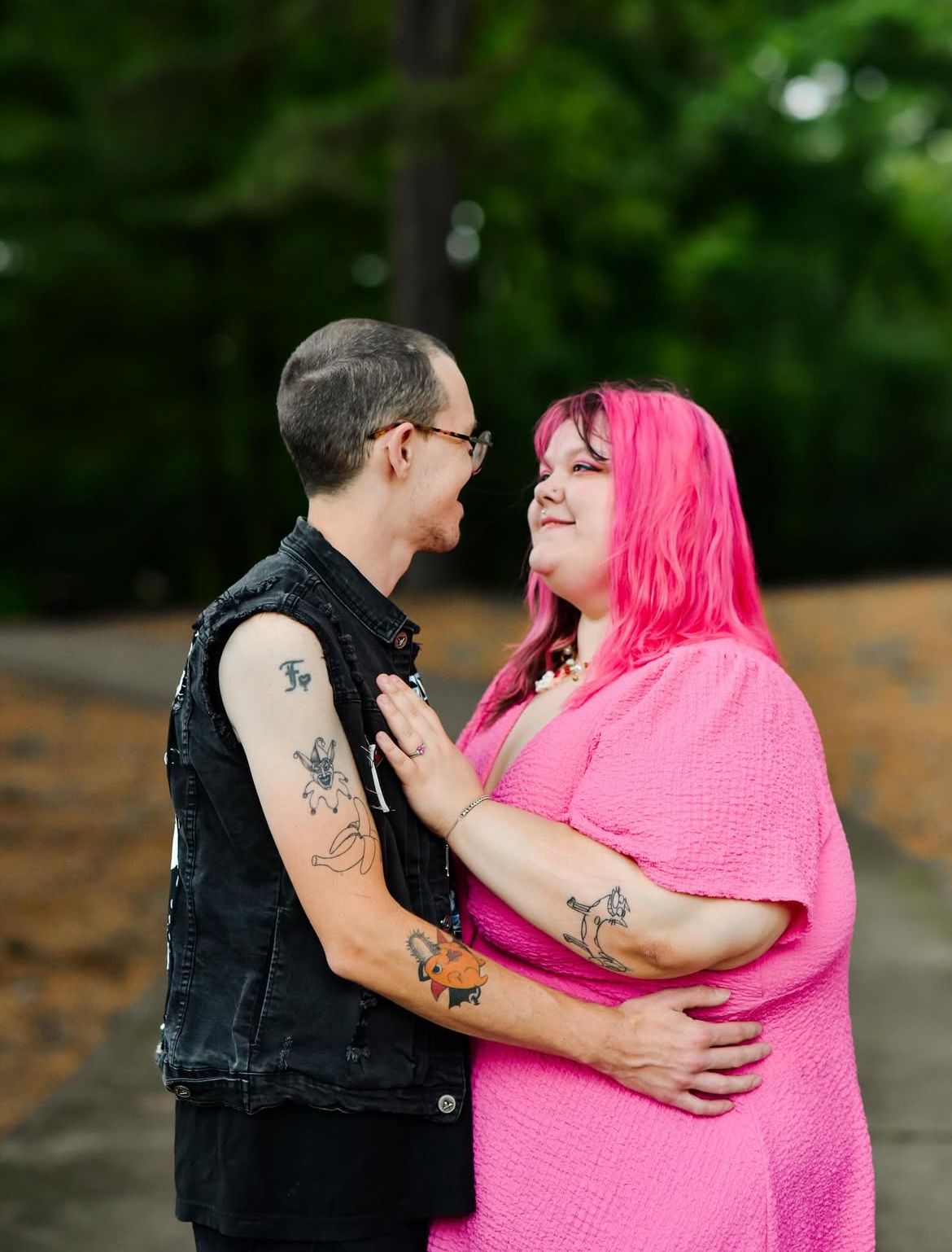 Couple embracing outdoors, woman with bright pink hair and pink dress, man with tattoos and denim vest, sharing a loving moment.