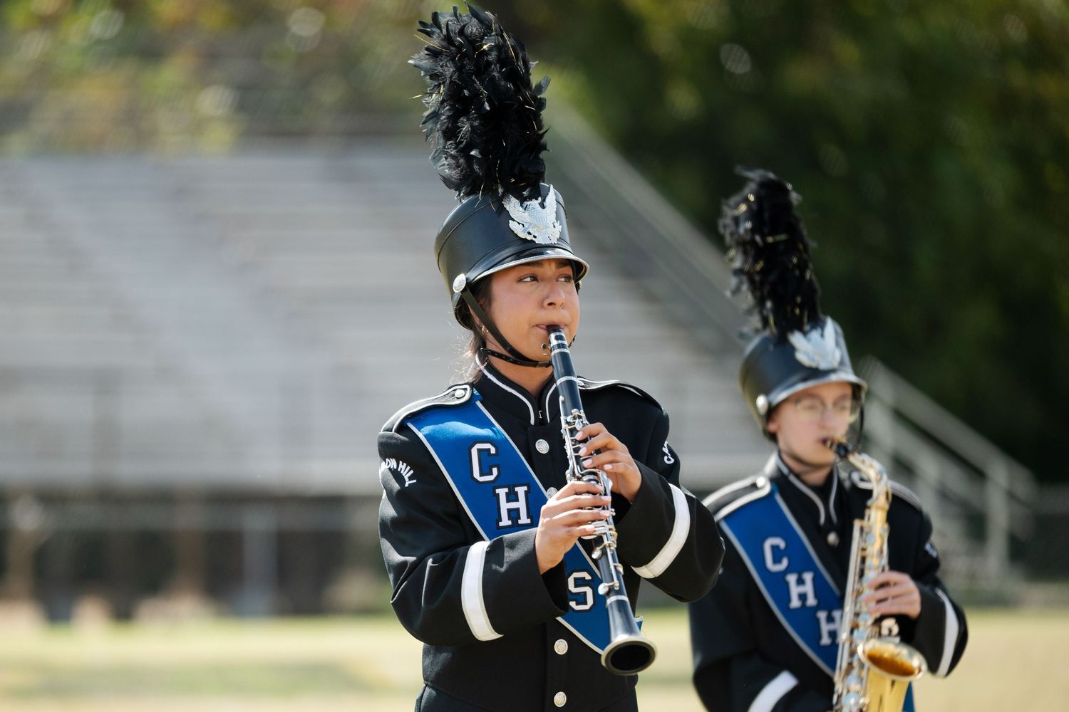 Marching band student playing clarinet in blue uniform with black feathered hat during outdoor performance.