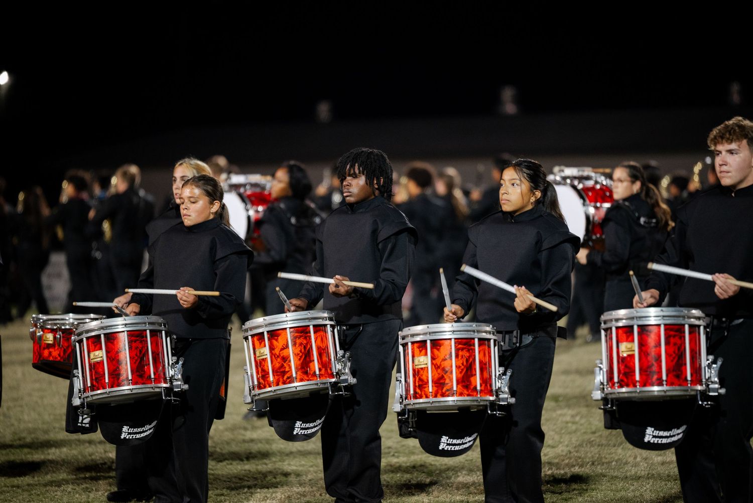 Marching band percussion members in black uniforms playing red snare drums at a night performance.