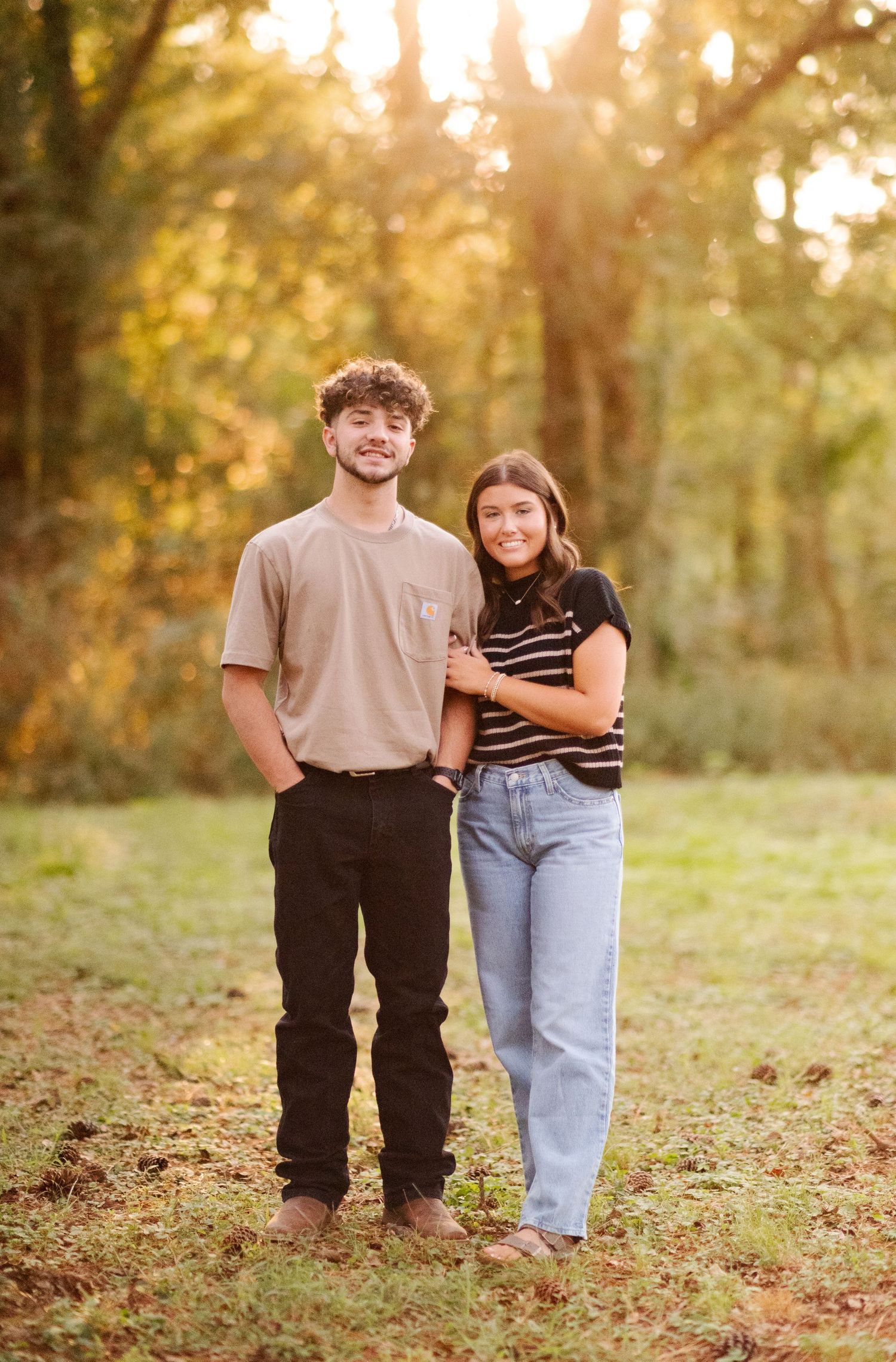 Smiling young couple standing together in a sunlit park with golden autumn trees behind them.