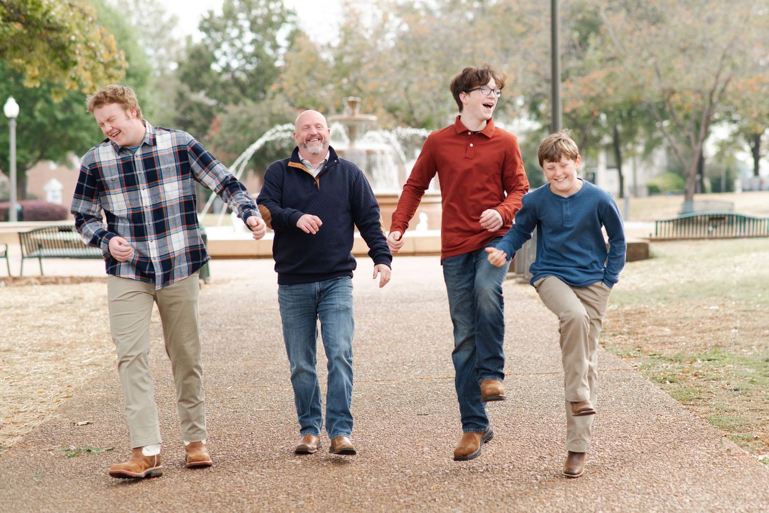 A father and three sons laugh and walk together along a park pathway with a fountain backdrop.