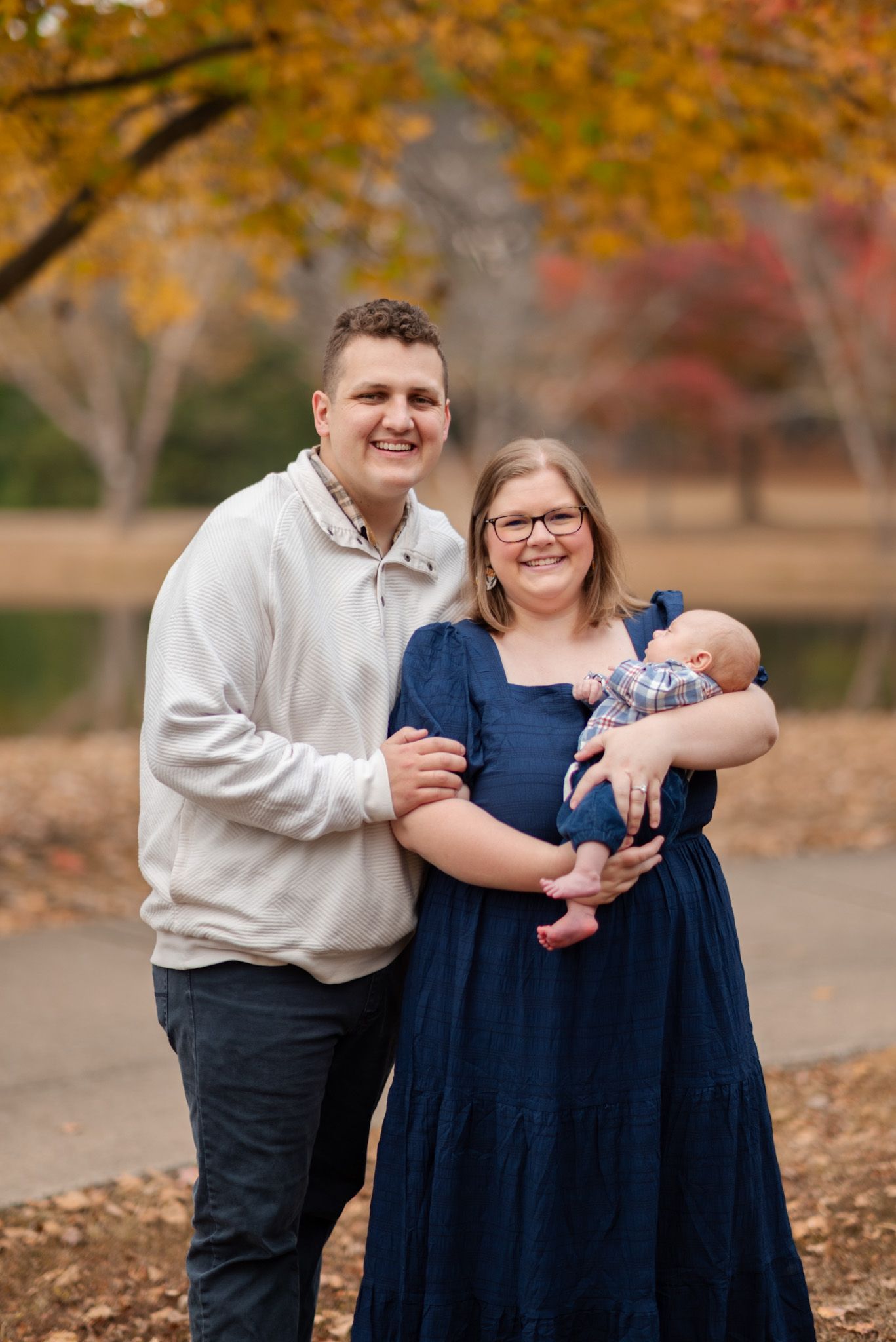 Smiling young family with newborn baby posing together outdoors under vibrant autumn foliage.