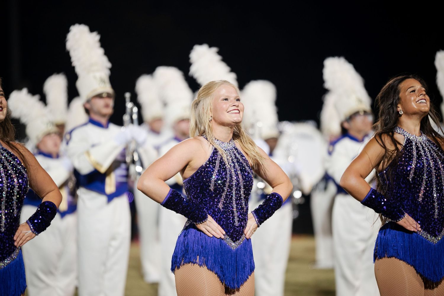 Smiling female dancer in blue sequin costume performs with marching band members in white uniforms.