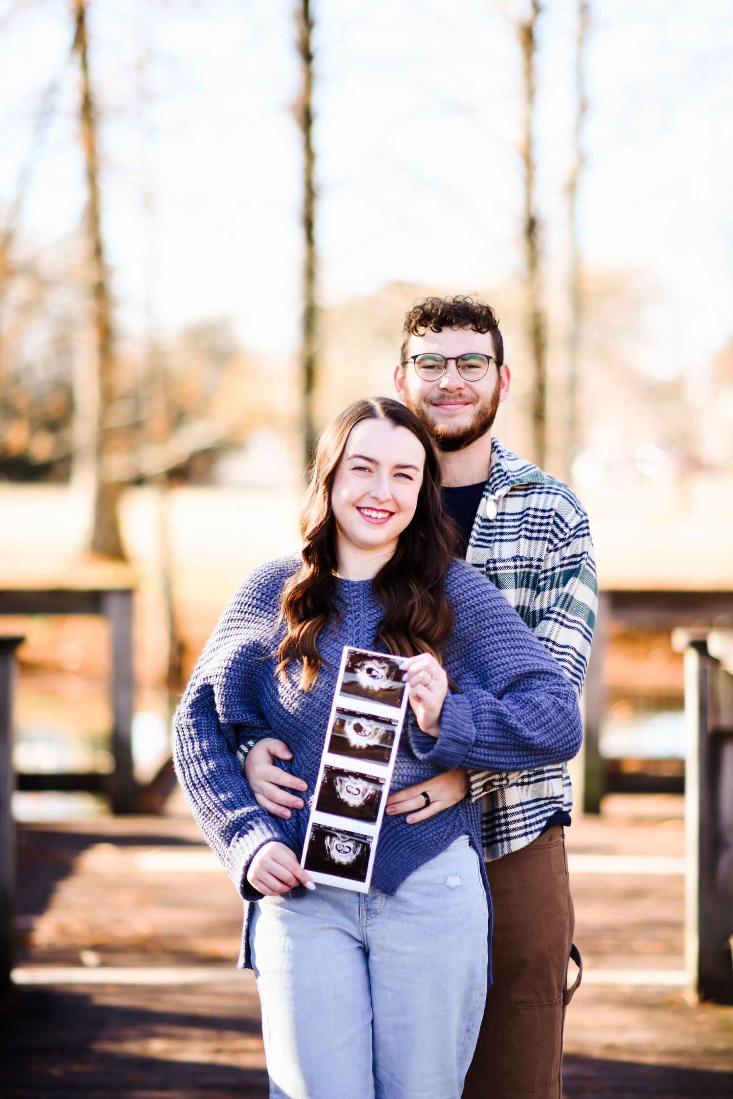 Happy couple holding ultrasound photos while embracing outdoors in sunlight.