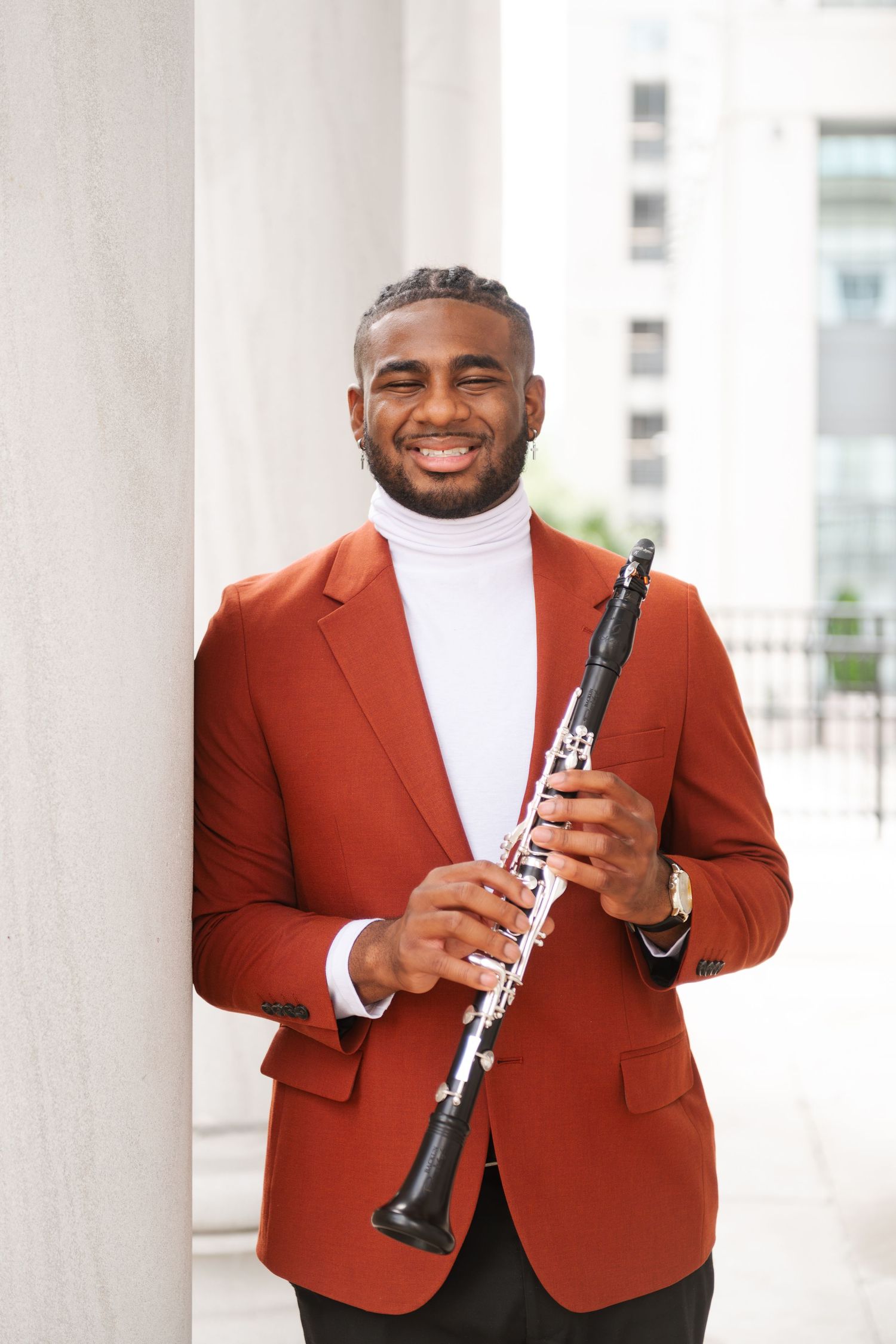 Smiling Black musician in red blazer holds clarinet outdoors near white columns.