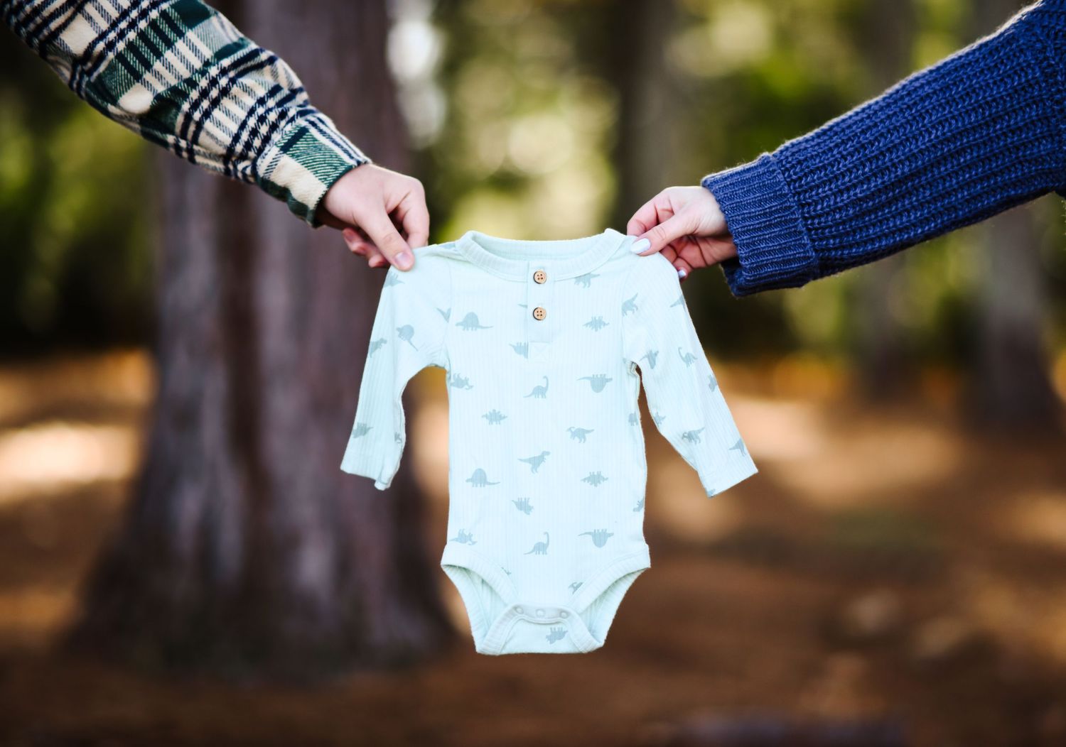 Two people holding a small dinosaur print baby onesie outdoors in the woods.