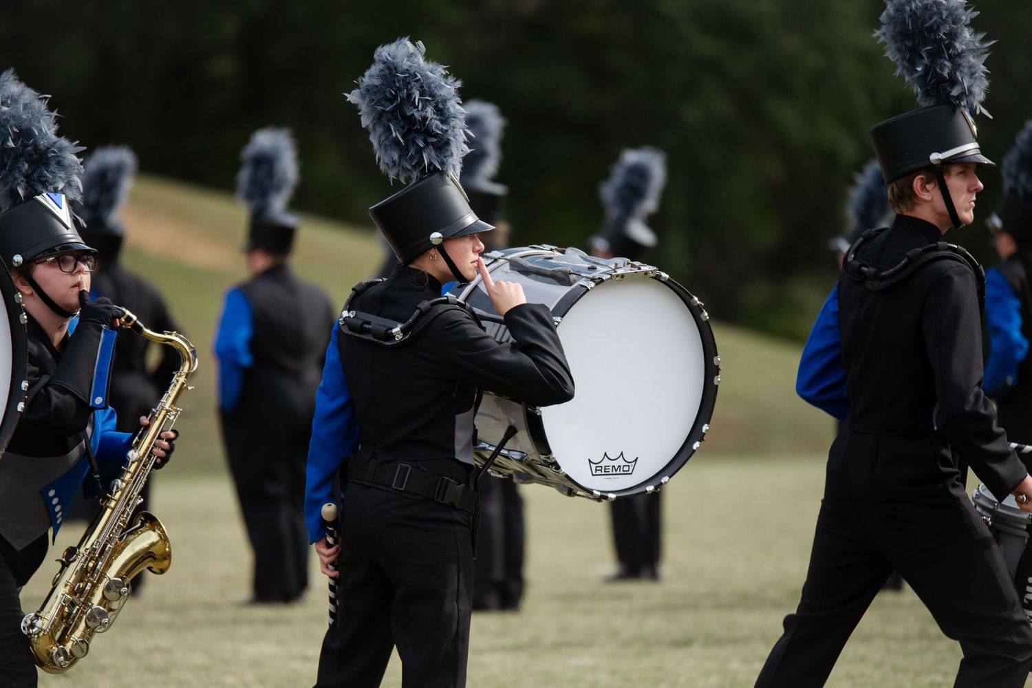 Marching band members in black uniforms play bass drum and saxophone on grassy field.