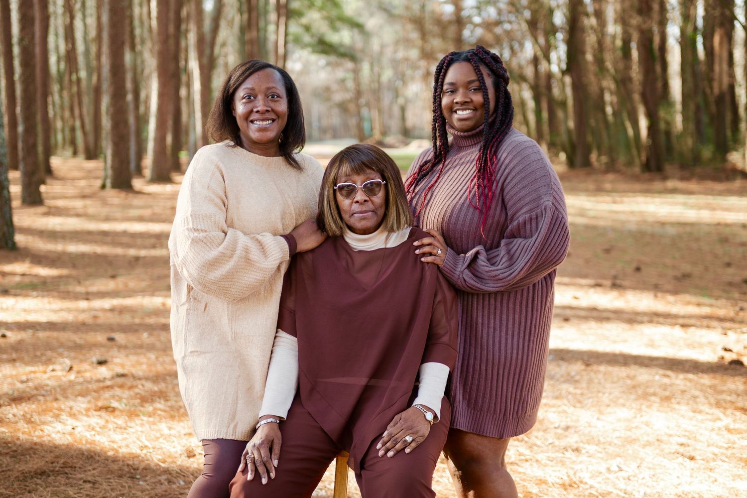 Three women smile together for portrait in sunglasses and earth-tone outfits in pine forest.