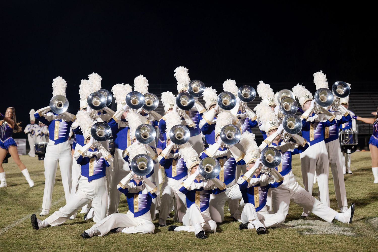 Marching band in white and blue uniforms poses with brass instruments at night.