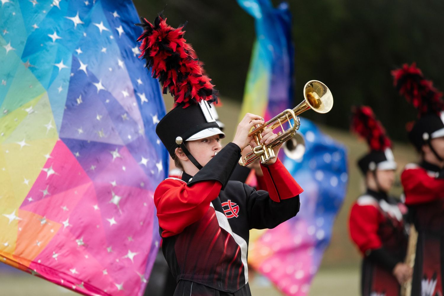 A marching band trumpet player performs beside a colorful rainbow star flag during an outdoor show.