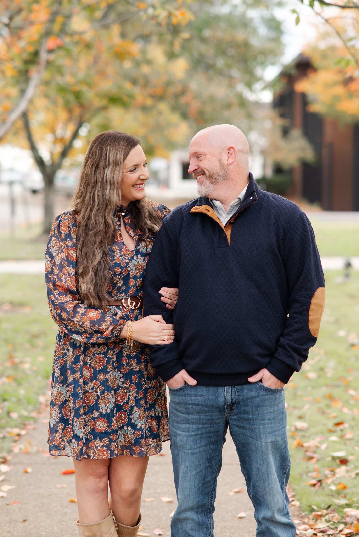 A smiling couple walks arm in arm on a leaf-covered path during fall engagement photos.