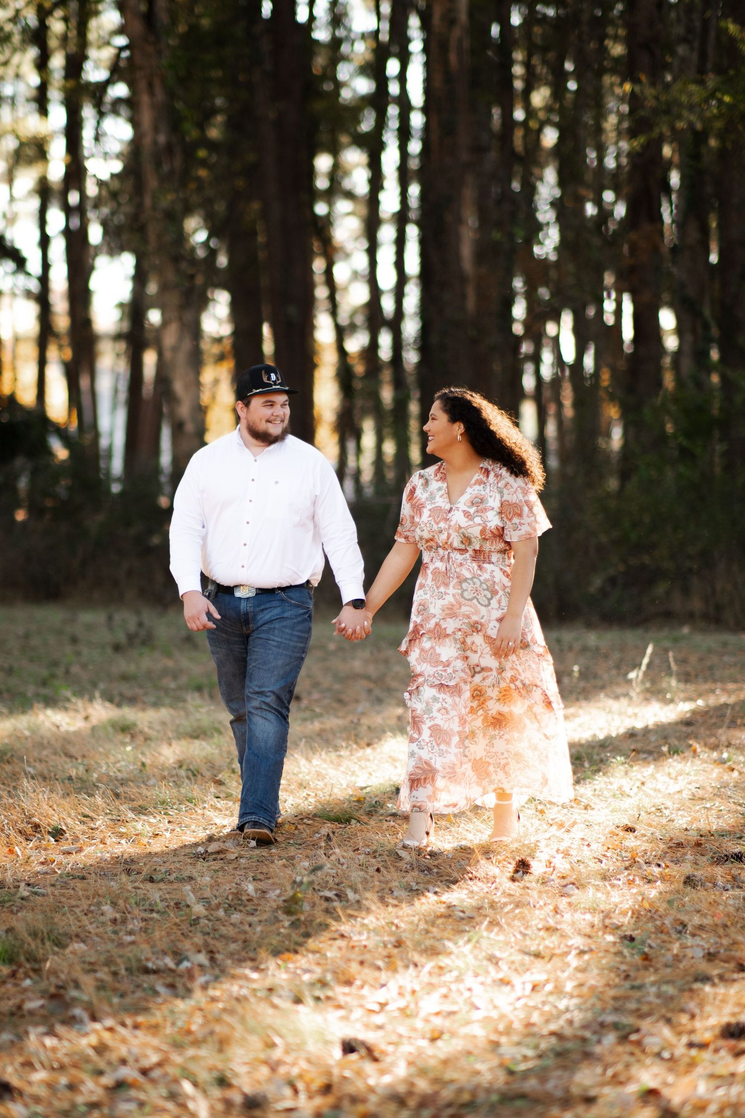 A couple holds hands and walks through a sunlit pine forest in autumn engagement photos.